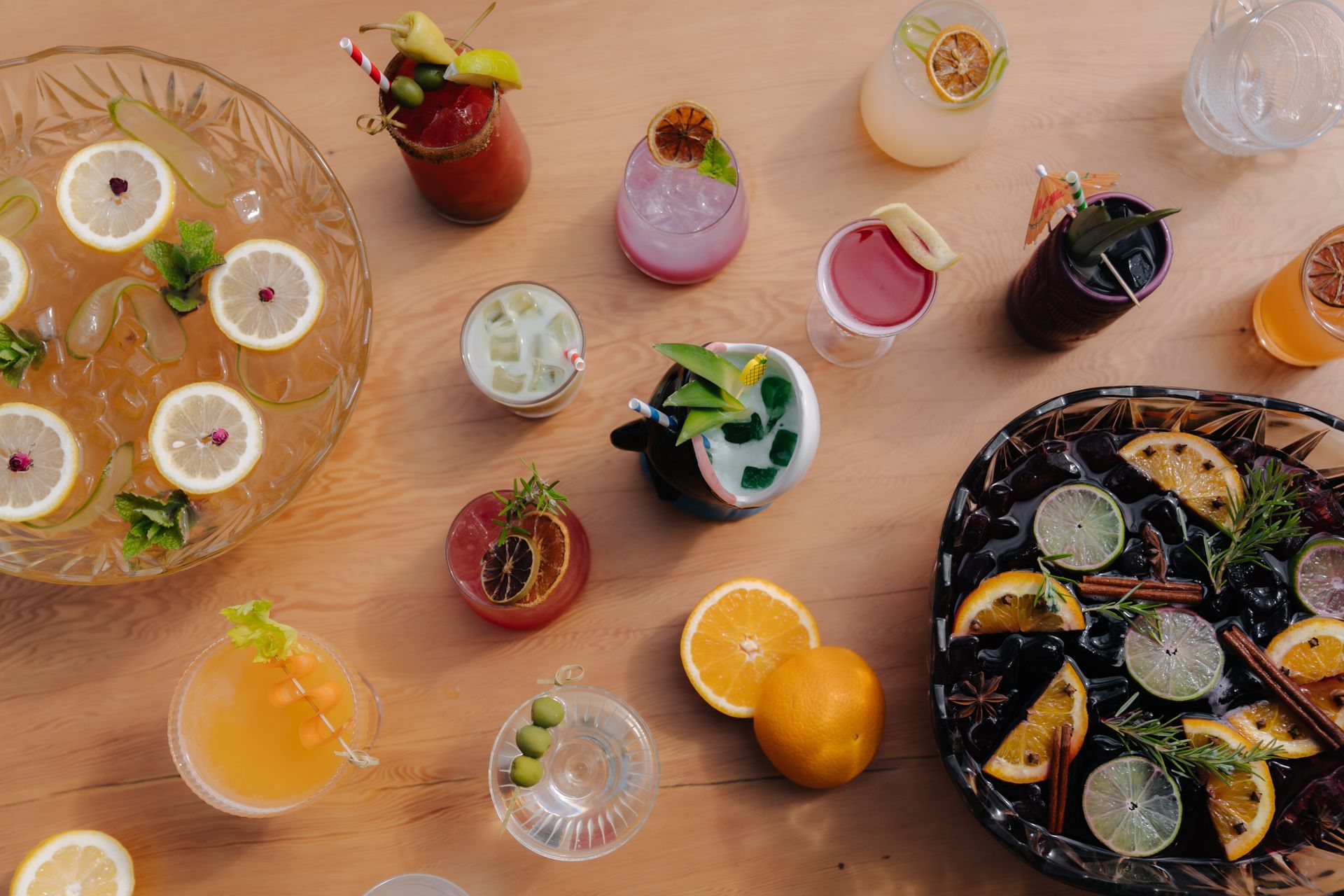 An assortment of colorful cocktails, garnishes, and two large punch bowls arranged on a light wooden table.