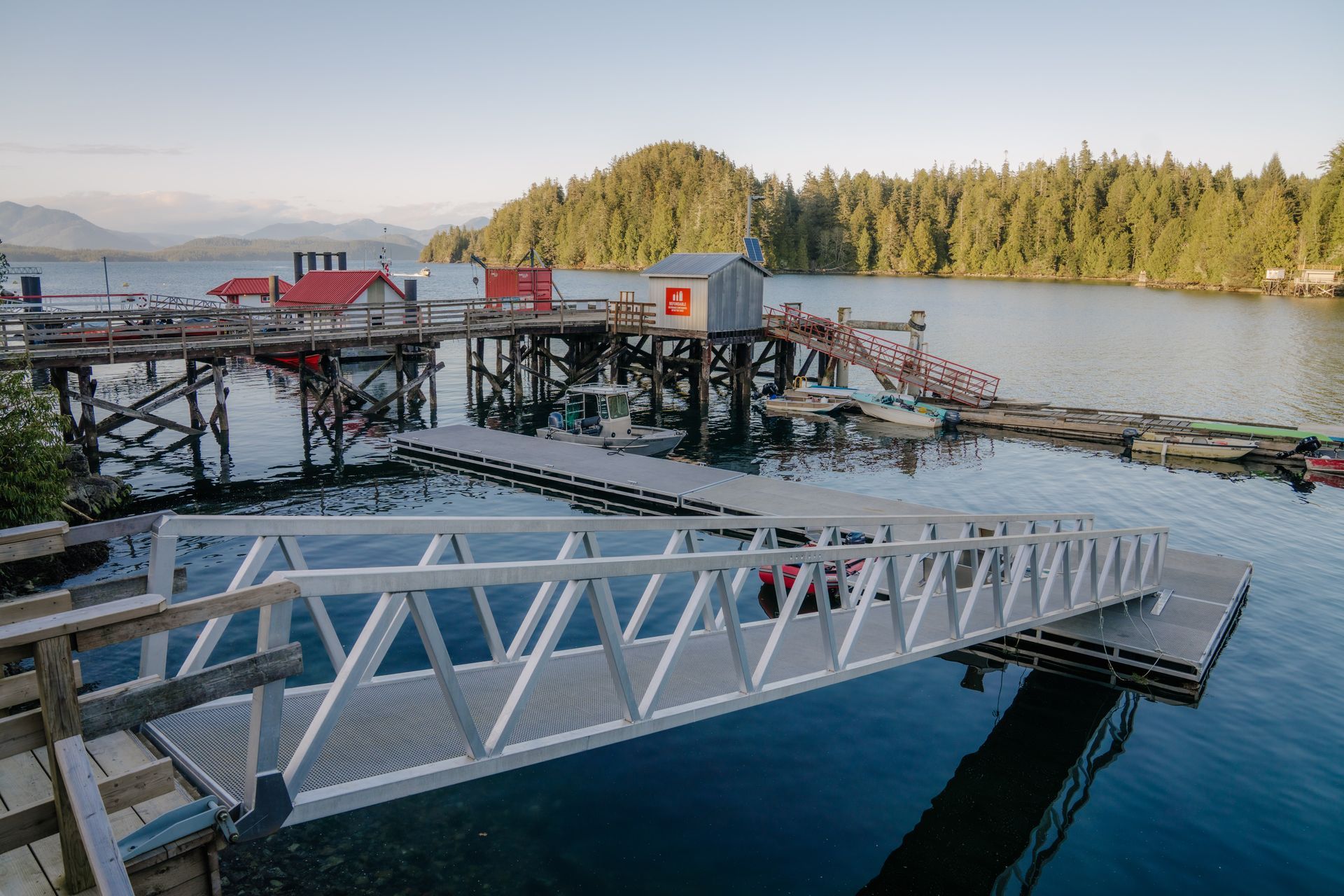 A long aluminum ramp leads to a floating dock in a calm bay, with wooden piers and forest-covered hills in the background.