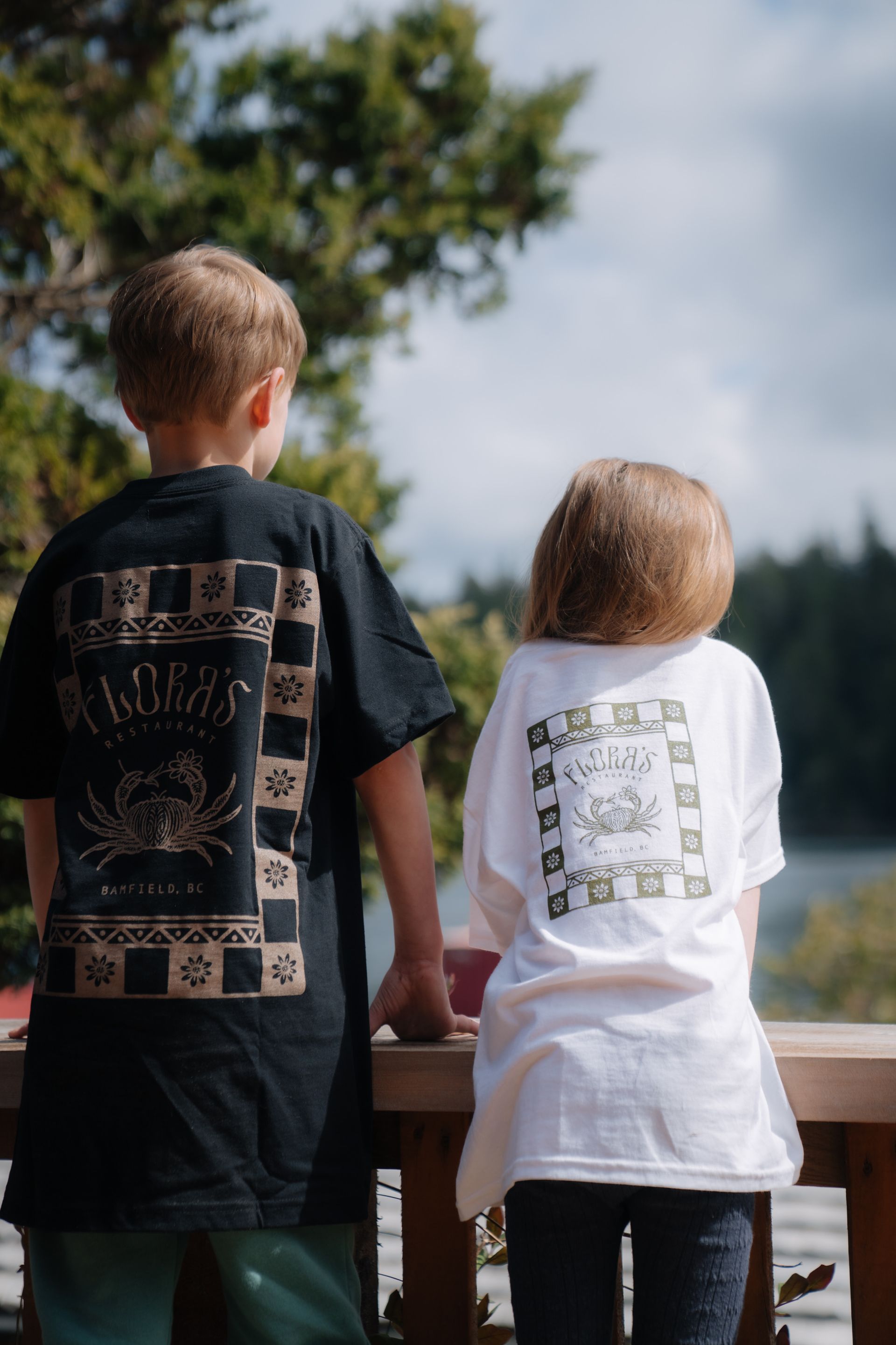 Two people stand side-by-side wearing black and white shirts with graphic crab designs, facing a view of trees and water.