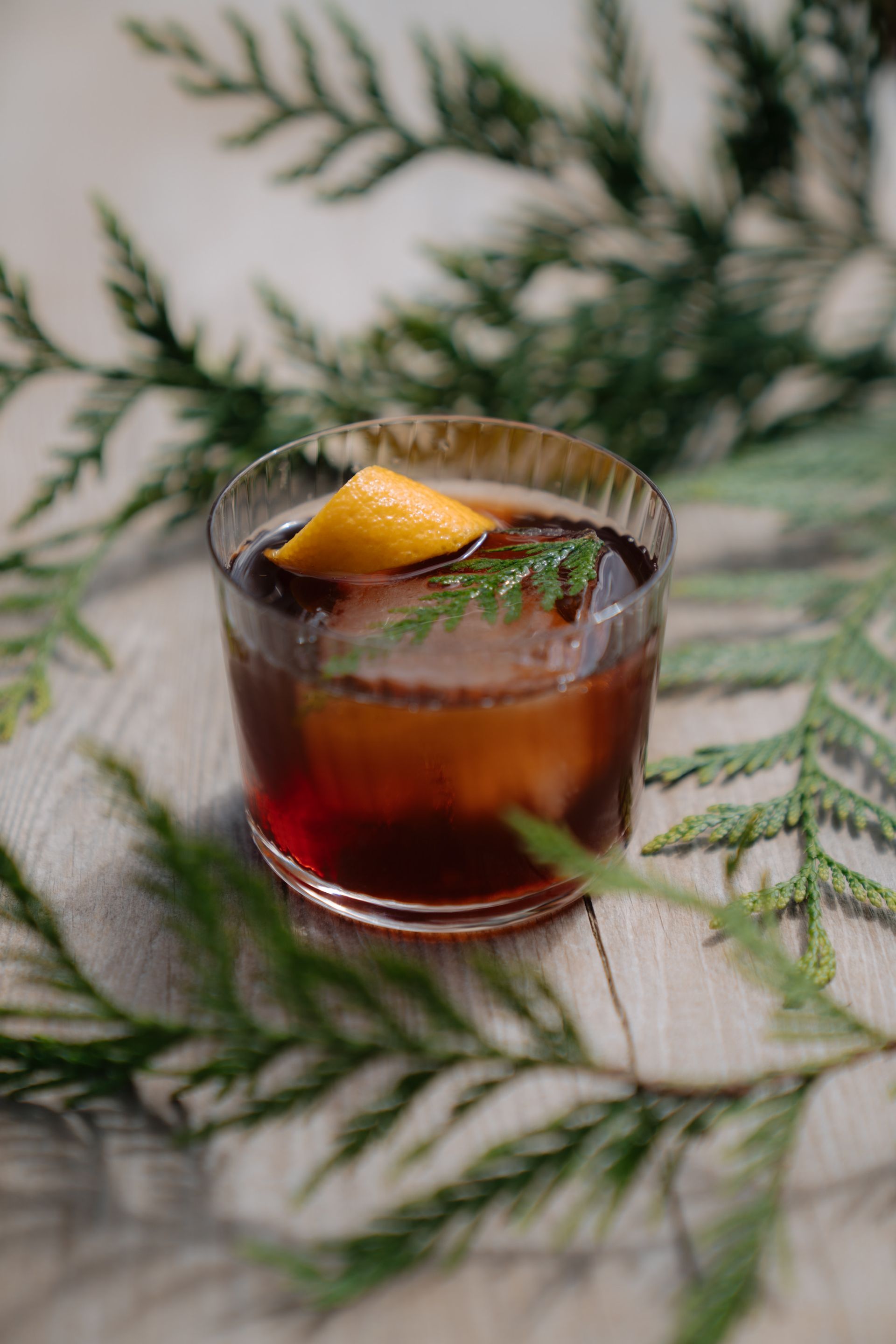 A cocktail with a large ice cube, garnished with an orange twist and evergreen sprig, set amidst pine branches.