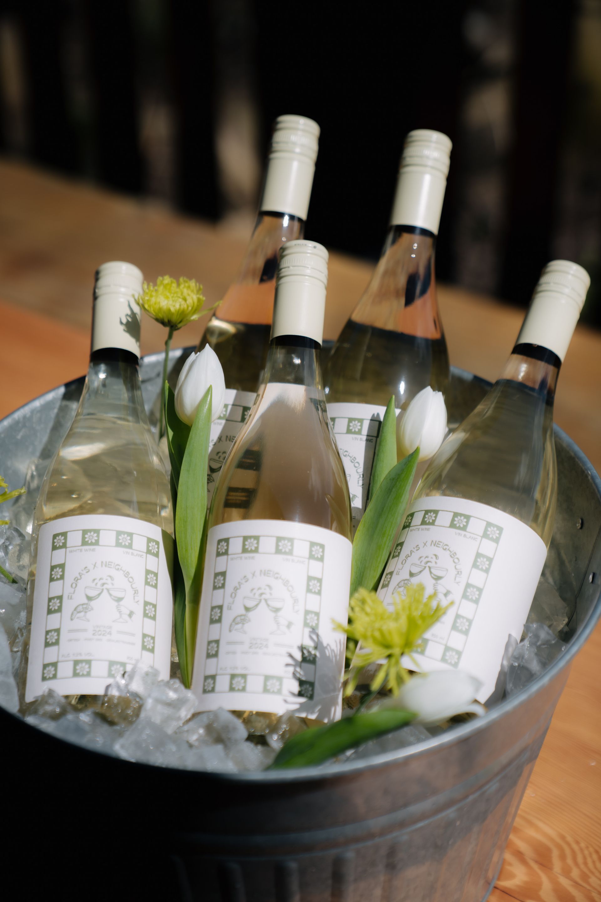Five bottles of wine chilled in a galvanized metal bucket filled with ice, accented with white tulips and green flowers.