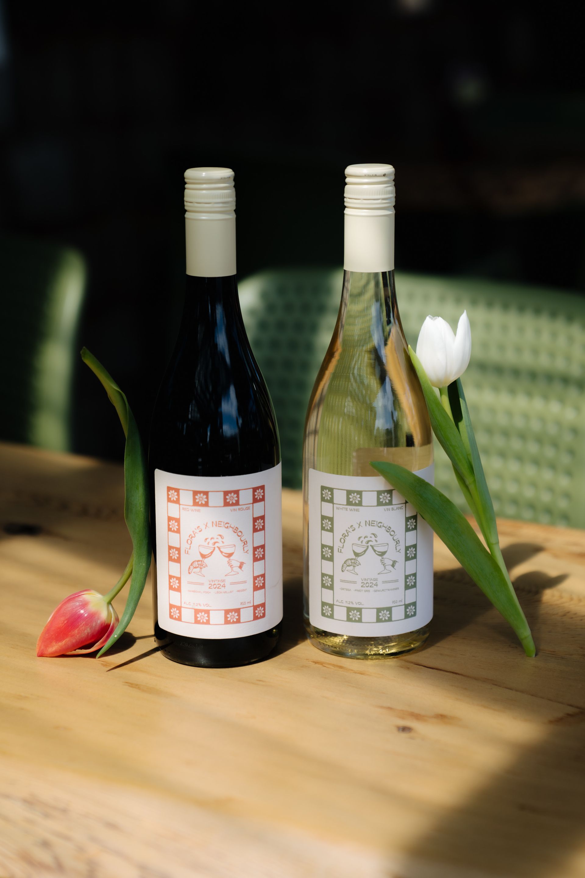 Two wine bottles, one red and one white, stand on a wooden table with a pink and a white tulip beside them.