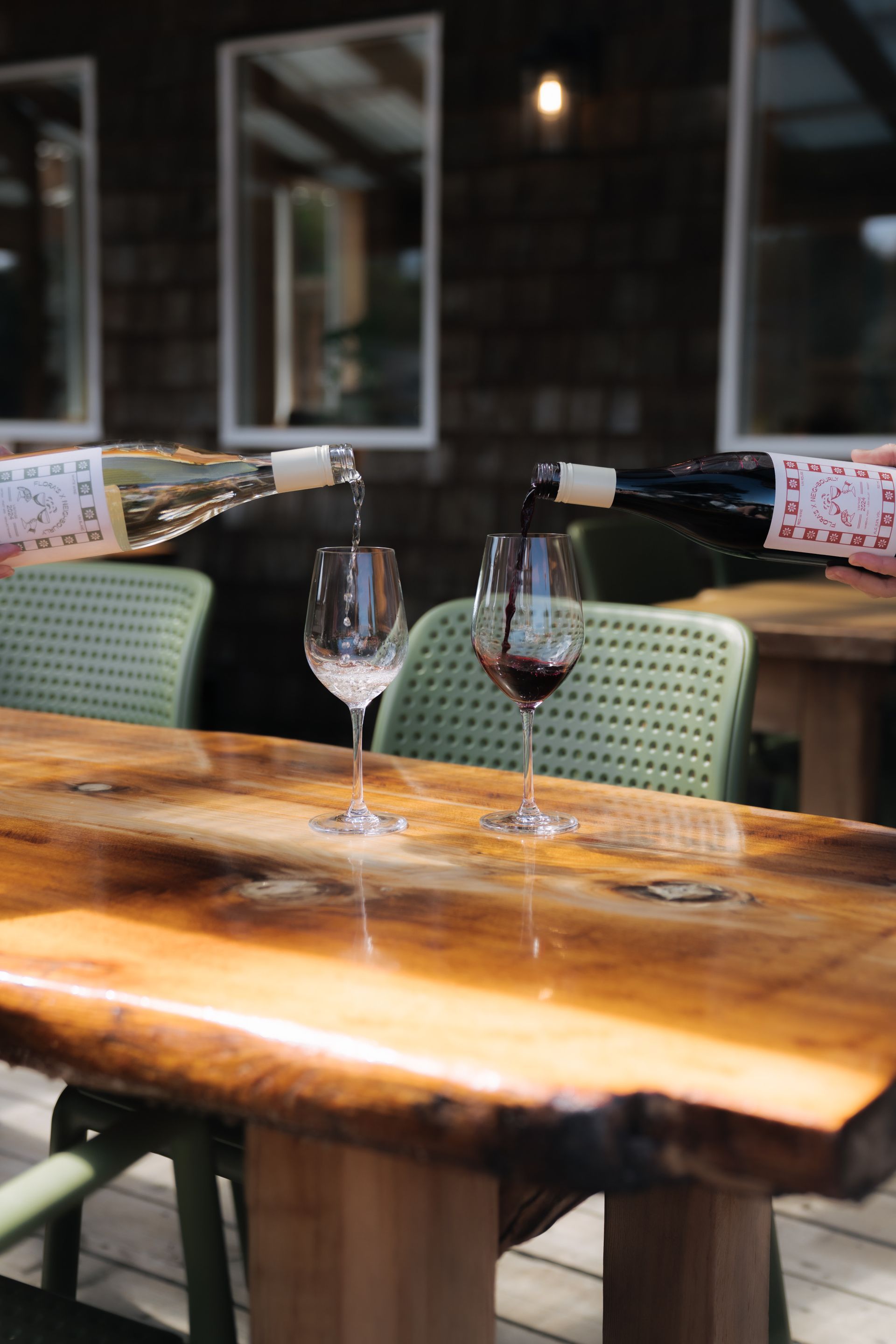 Two bottles of wine are poured simultaneously into separate glasses on a rustic wooden table.