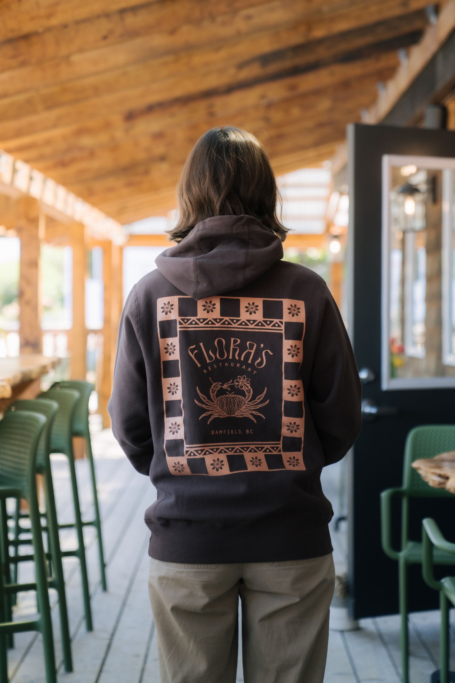 A person wears a dark brown hoodie with a rectangular graphic featuring a crab illustration on an outdoor patio.