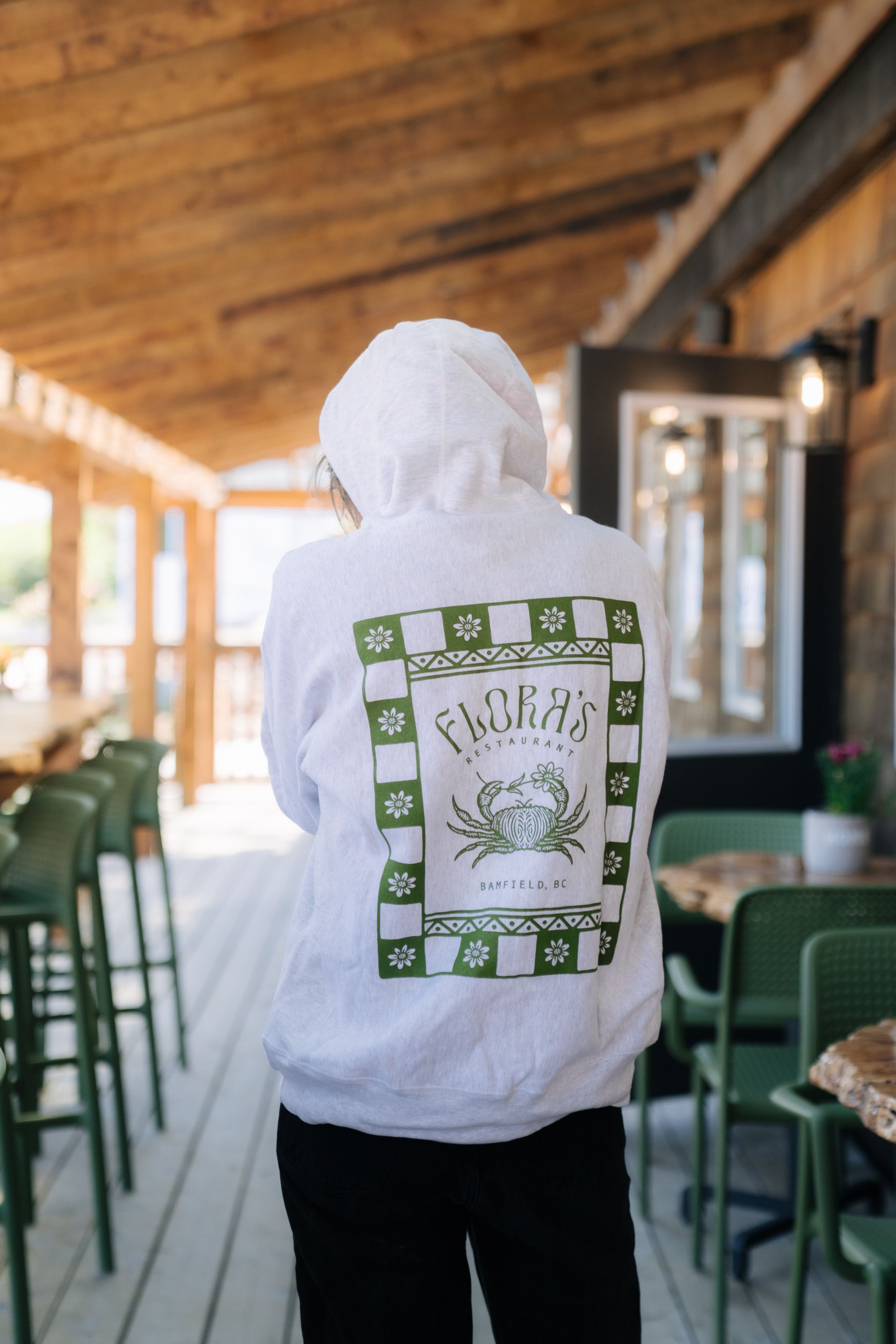 A person wearing a white hooded sweatshirt with a green graphic of a crab, seen from behind on an outdoor patio.