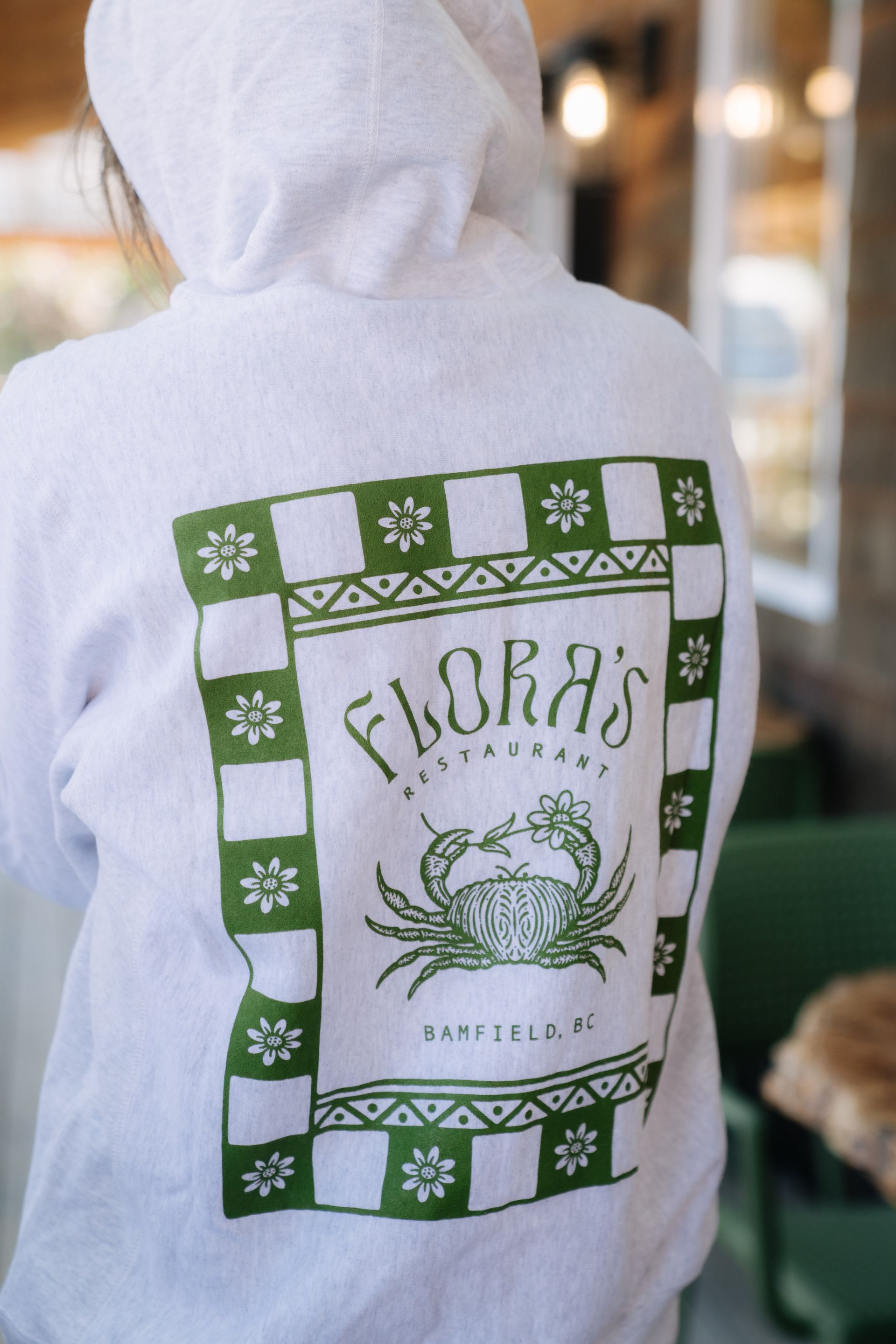 A white hoodie viewed from behind, featuring a green square graphic with a crab, floral accents, and stylized text.