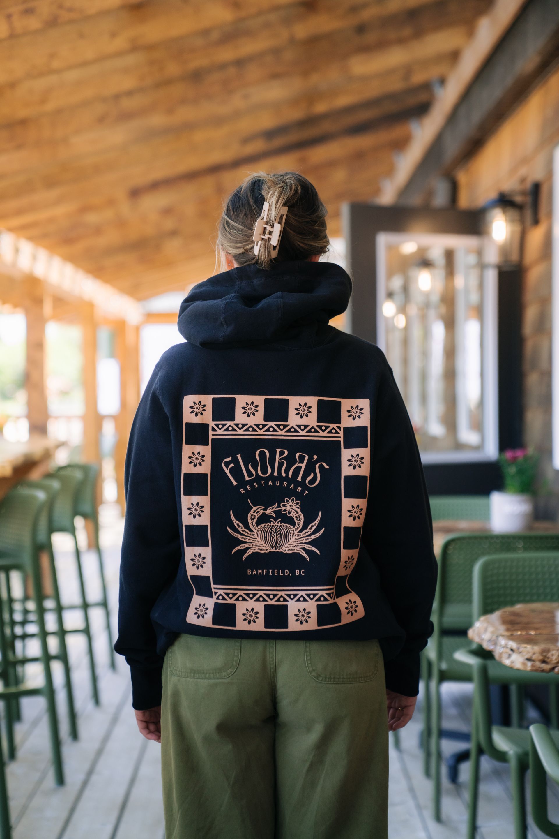 A person stands wearing a black hoodie with a tan, graphic-printed square on the back, inside a rustic, outdoor eatery.