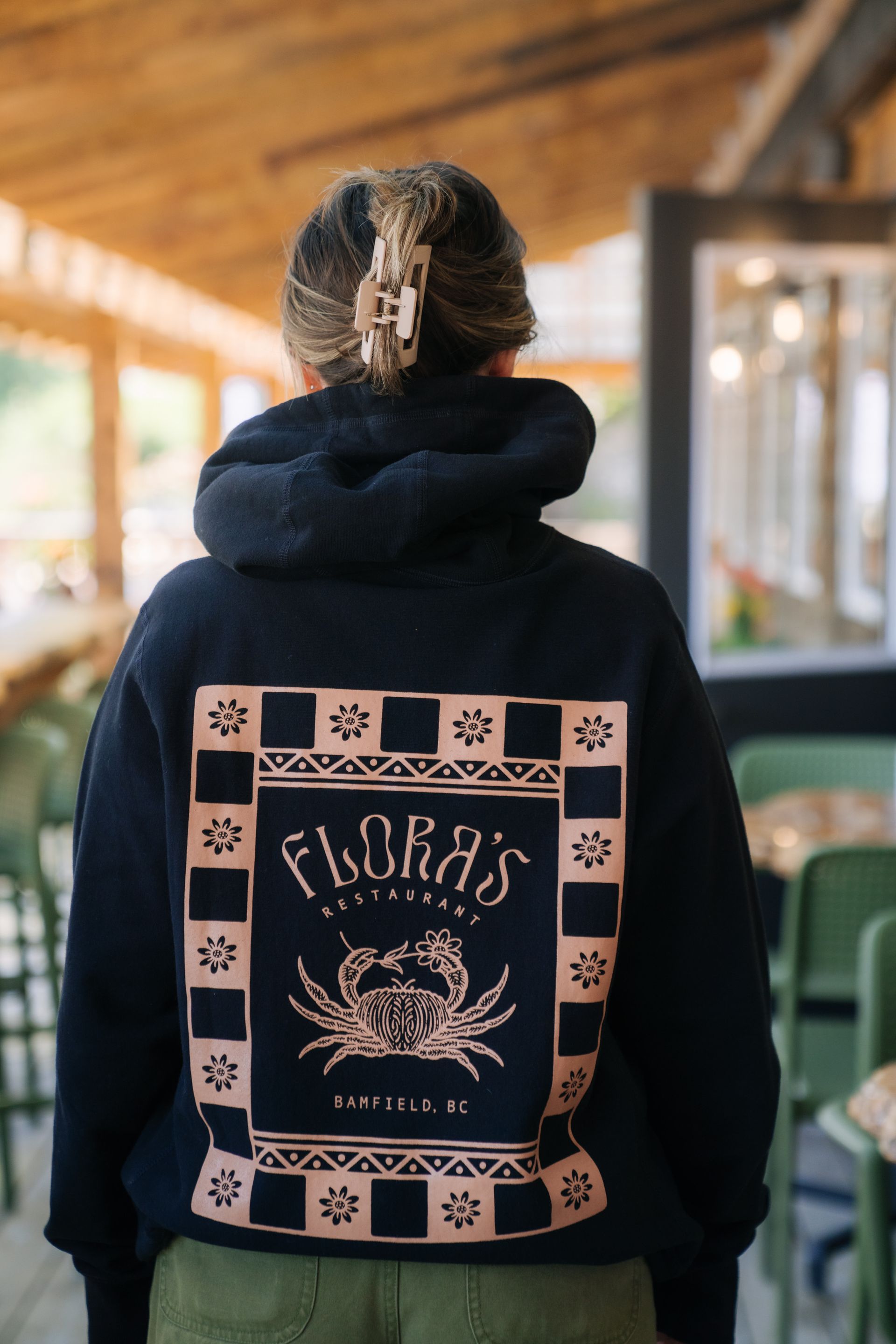 A person wears a black hoodie with a tan, block-print graphic on the back featuring a crab and 