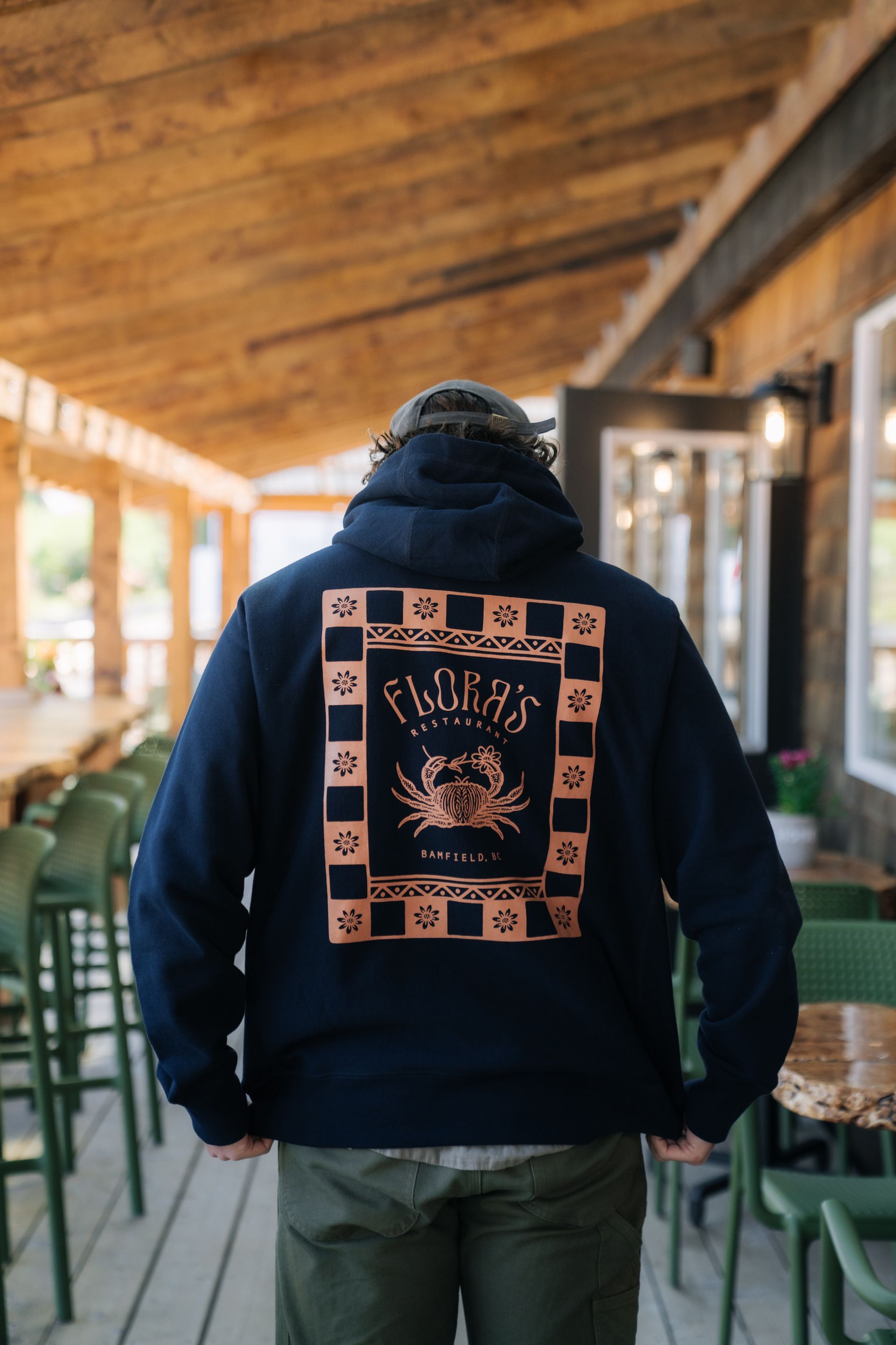 A person wearing a dark blue hoodie with a crab graphic on the back stands on a wooden outdoor deck.