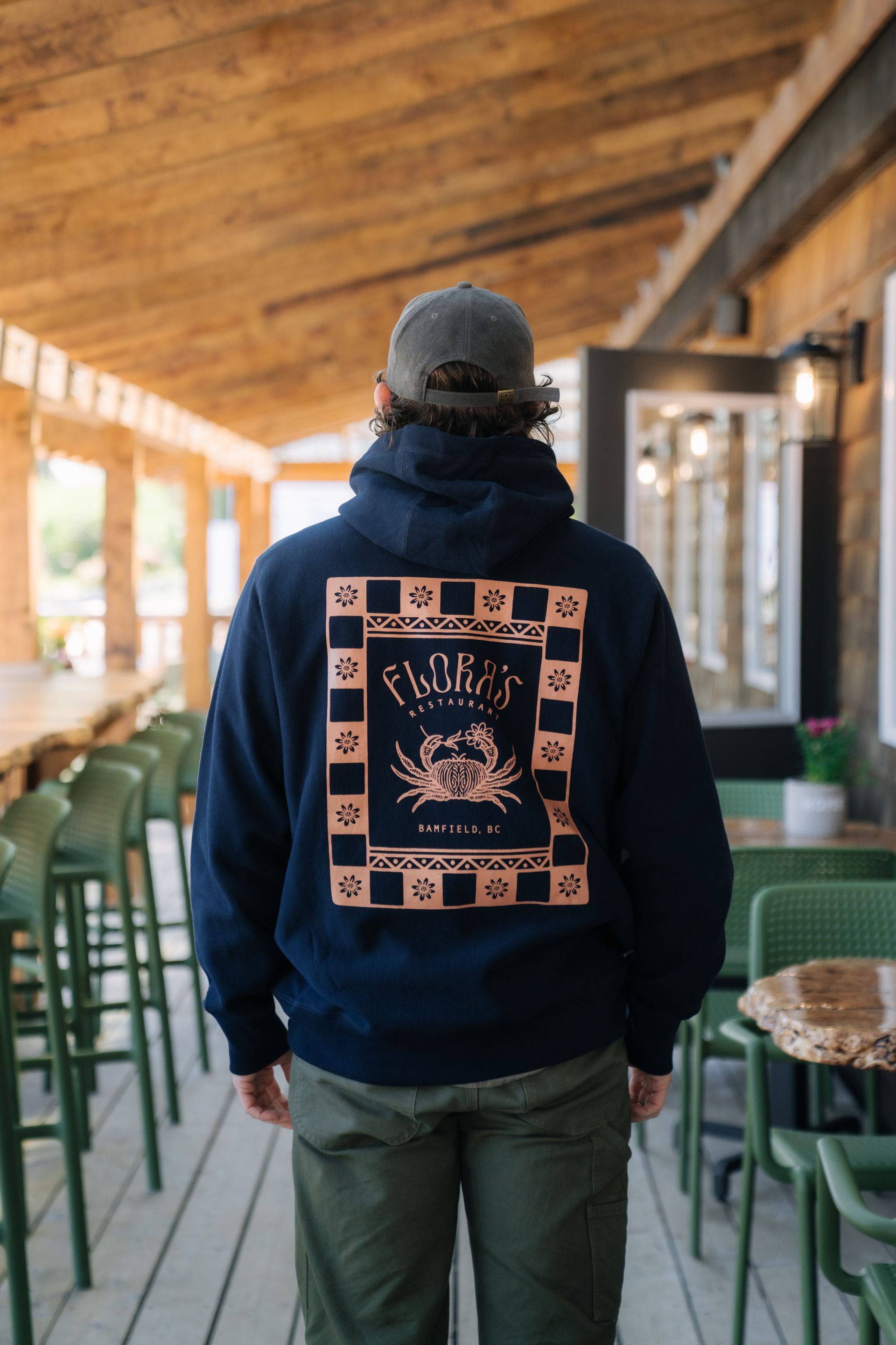 A person wears a navy hoodie with a crab graphic on the back, standing on a wooden deck by outdoor tables.