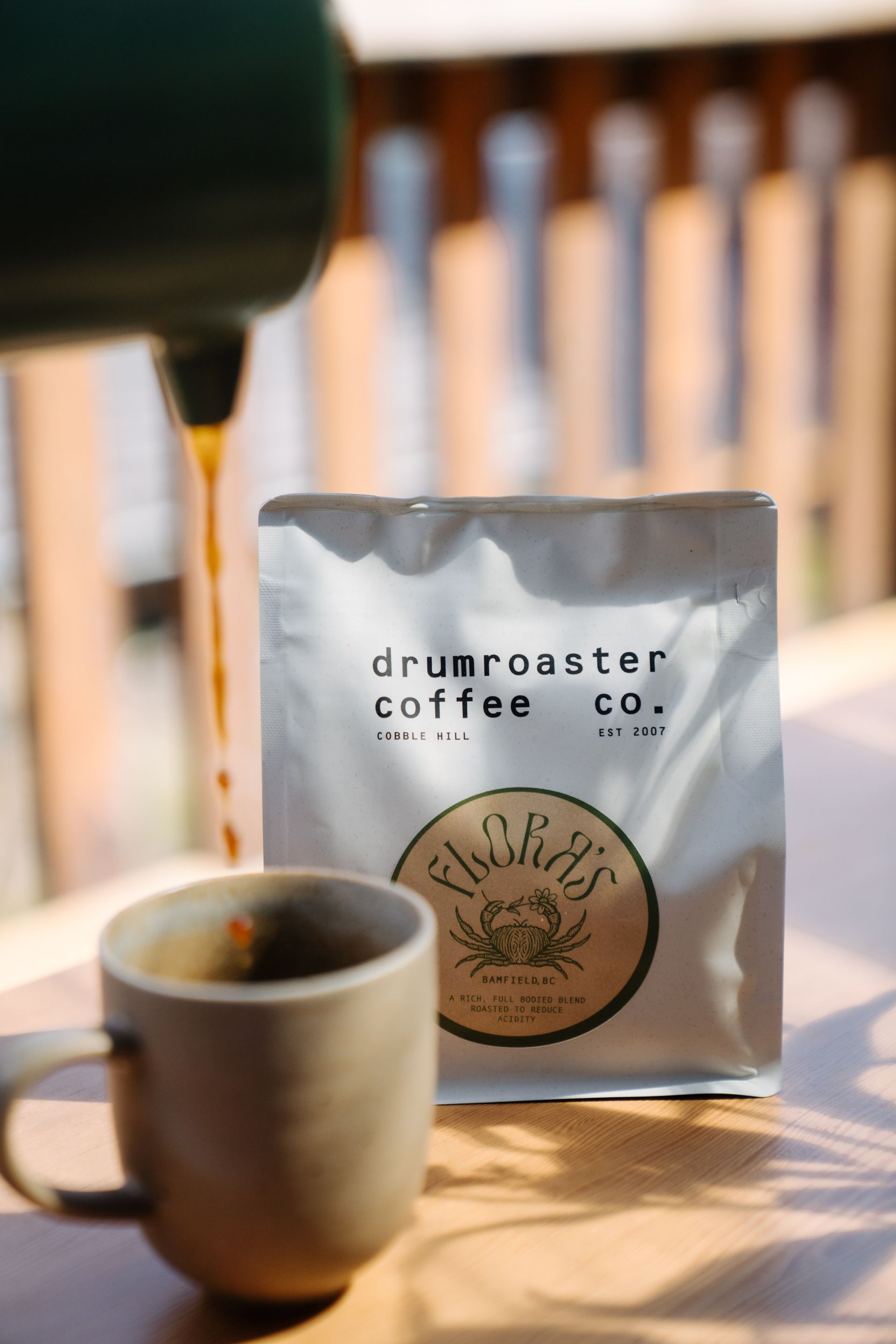 A bag of Drumroaster Coffee beside a tan mug as dark coffee pours from a brewer on a sunlit table.
