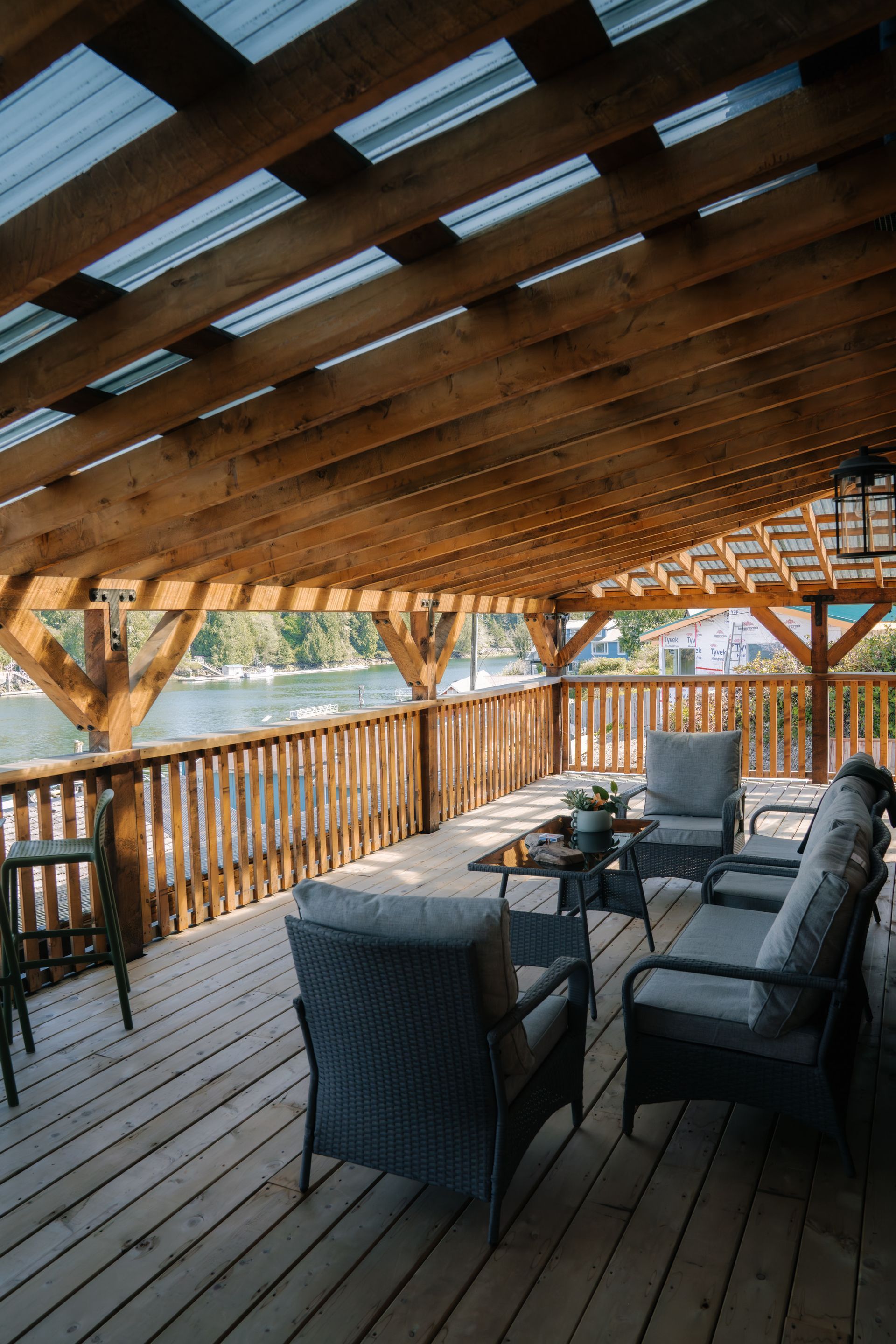 A wooden deck with a covered pergola featuring outdoor furniture, overlooking a body of water with trees in the distance.