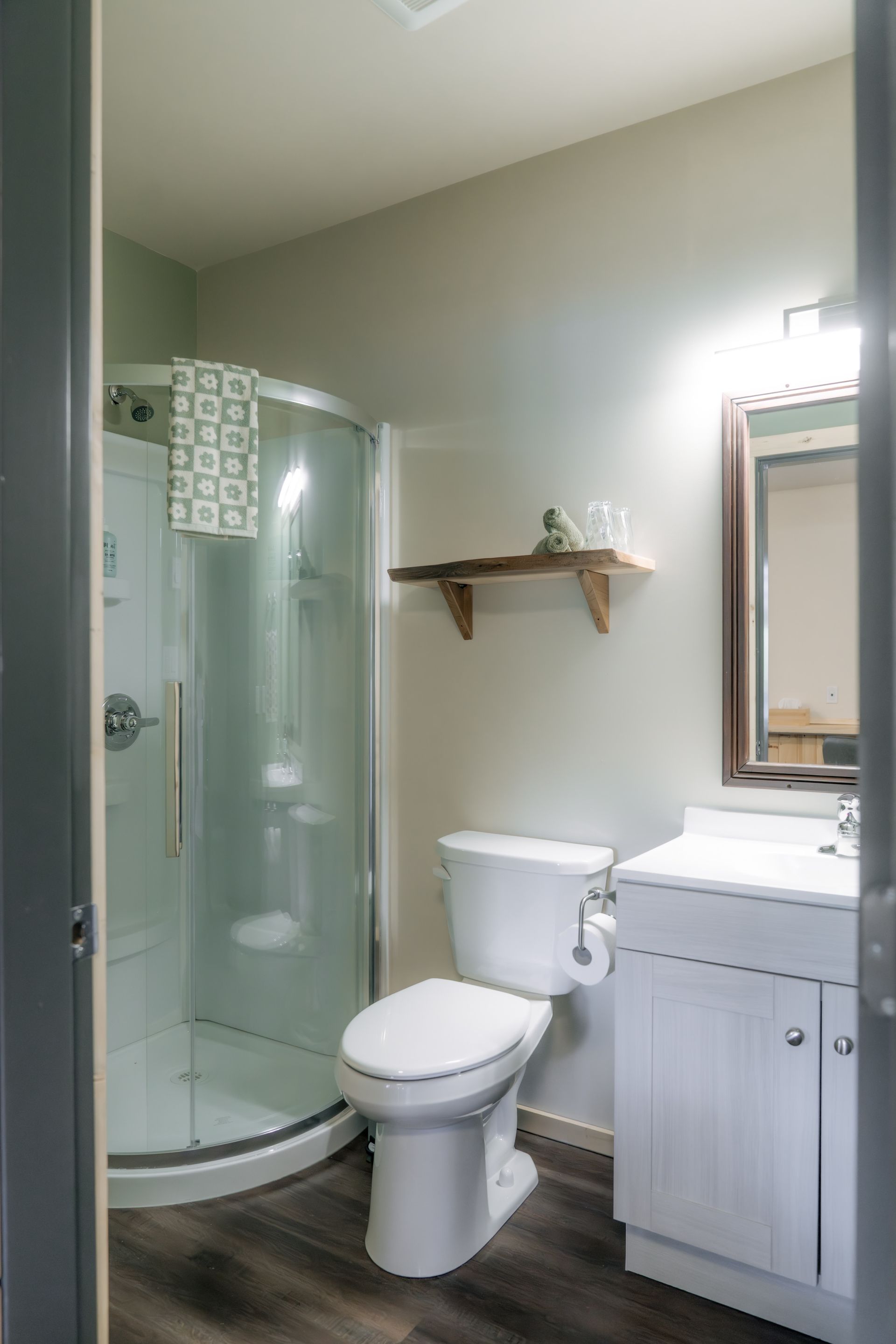 A small, bright bathroom with a curved glass shower, white toilet, floating wooden shelf, and a white vanity with mirror.