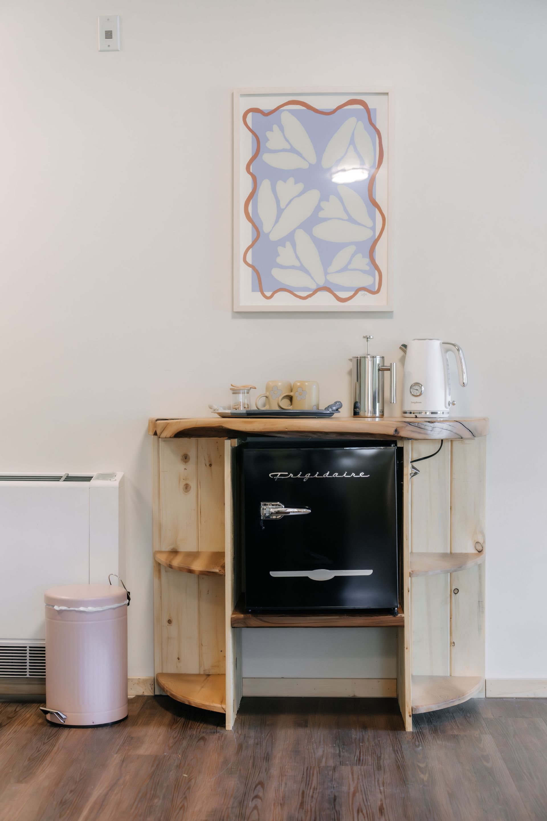 A light-wood coffee bar with a black mini-fridge, kettle, and wall art, next to a pink trash bin in a room.