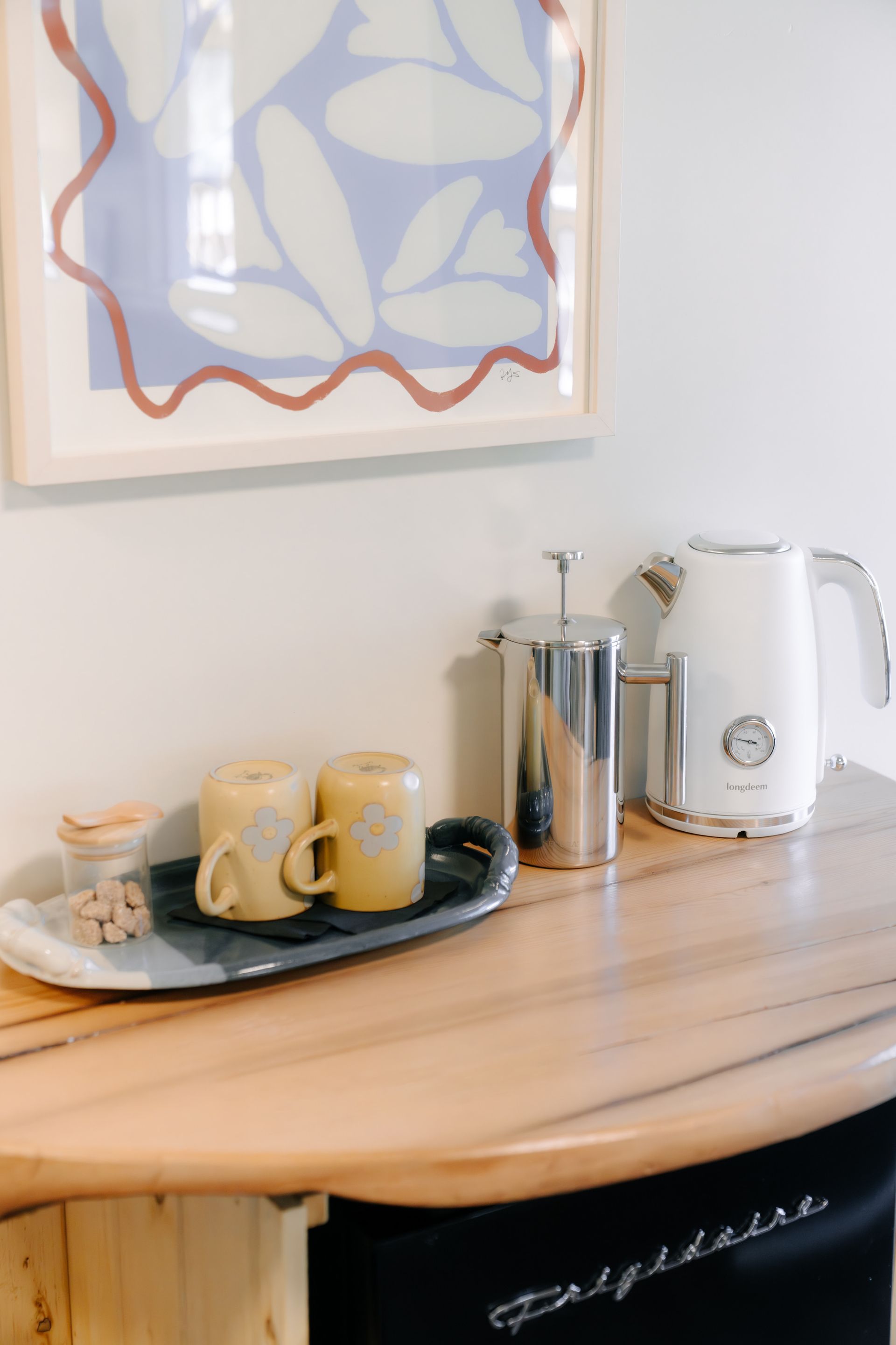 A wooden counter holds a white electric kettle, a metal French press, two yellow mugs on a tray, and a small jar of nuts.