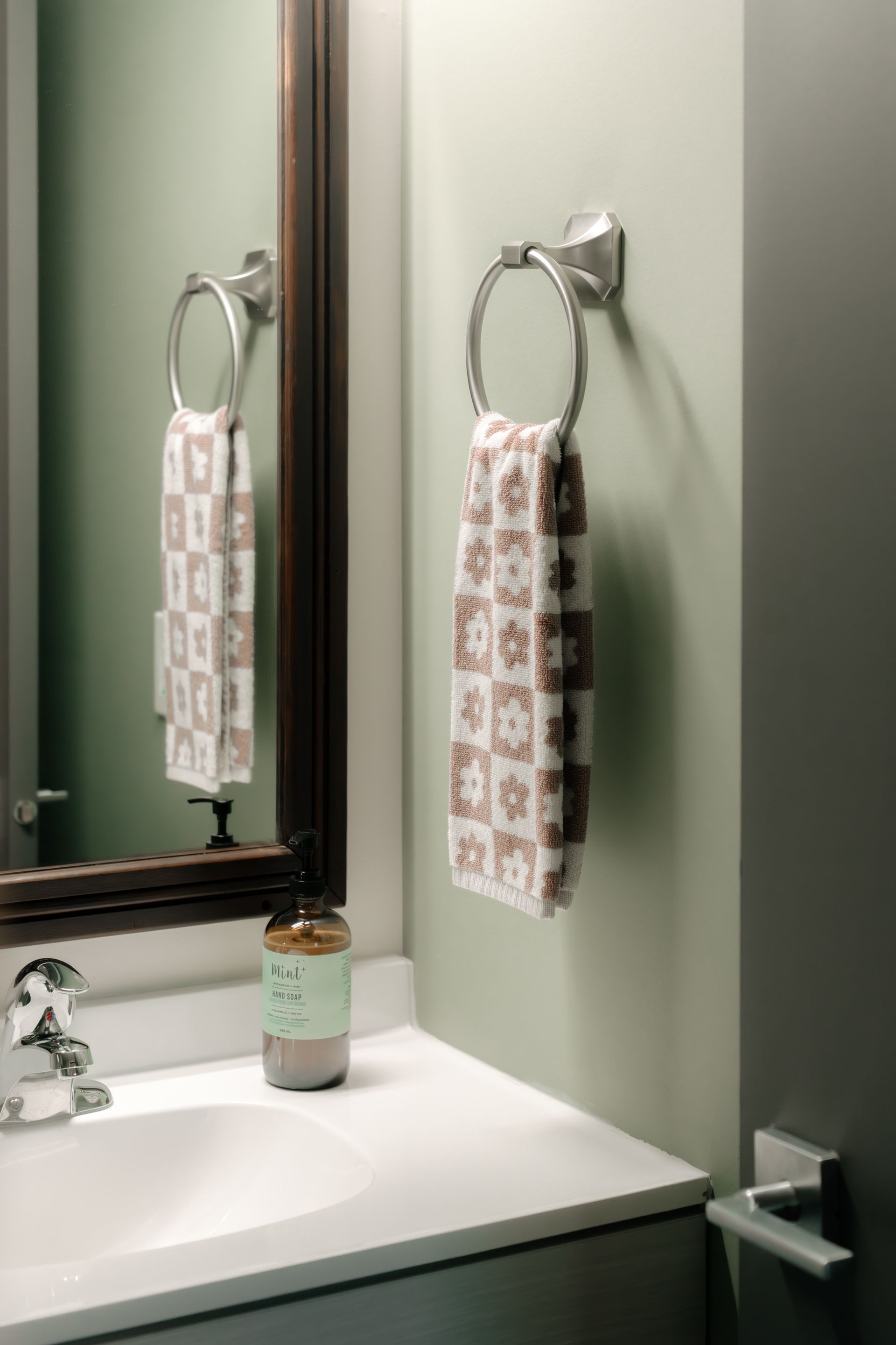 A sage green bathroom with a patterned hand towel hanging on a circular chrome ring beside a dark-framed mirror and sink.