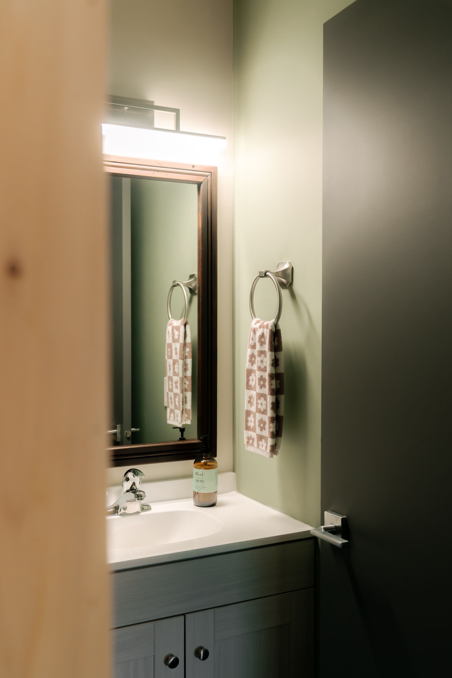 A bathroom vanity with a white sink, mirror, and towel ring on sage green walls next to a dark gray door.
