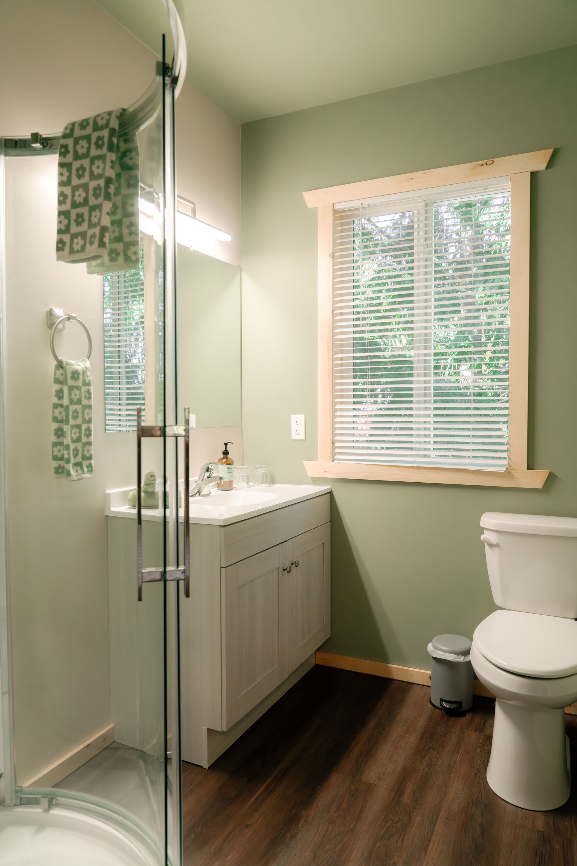 A bathroom with sage green walls, a glass-enclosed shower, a white vanity, a window with blinds, and a white toilet.