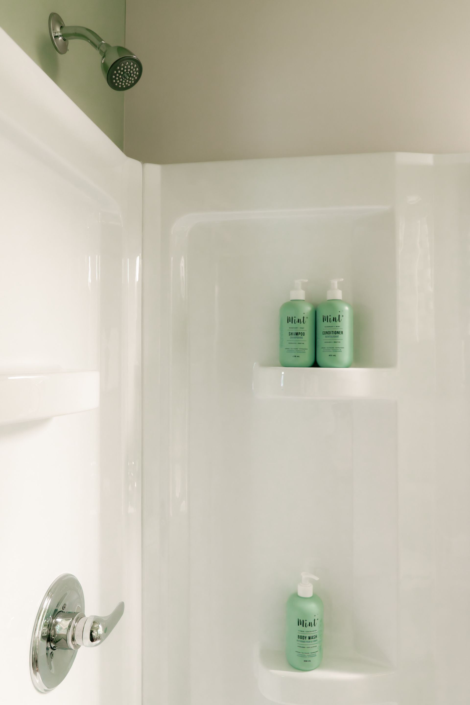 White shower stall with a chrome shower head and handle, featuring three light green bottles on built-in shelves.