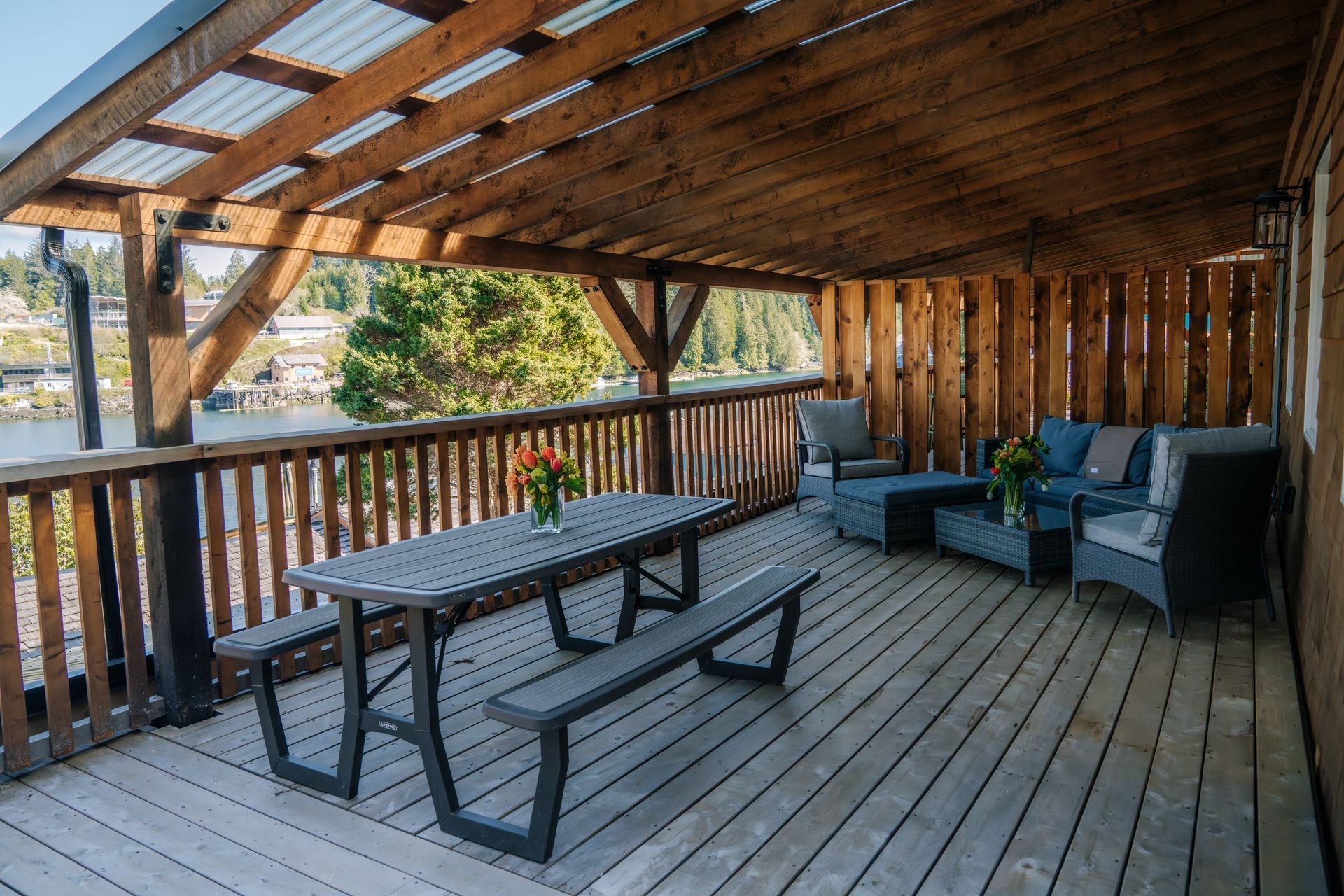 A covered wooden deck featuring a grey picnic table with benches and a separate lounge area with dark cushioned furniture.