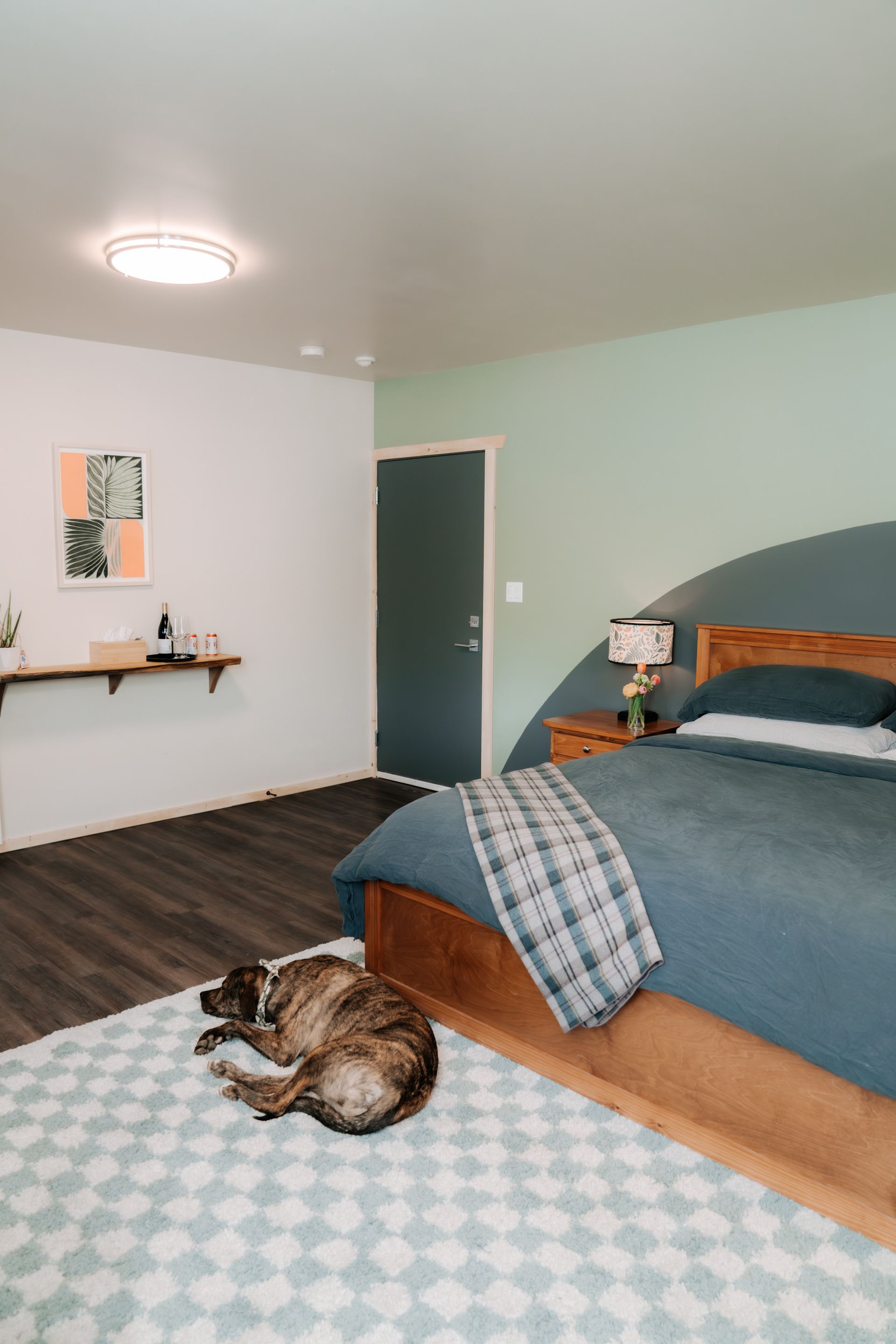 A bedroom with a dog sleeping on a patterned rug near a bed with blue bedding, a dark wood frame, and a painted headboard.