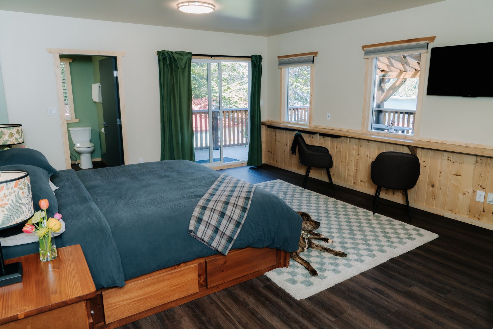A bedroom with a blue-covered bed, a rug, a dog resting on the floor, a desk area with two stools, and a small bathroom.