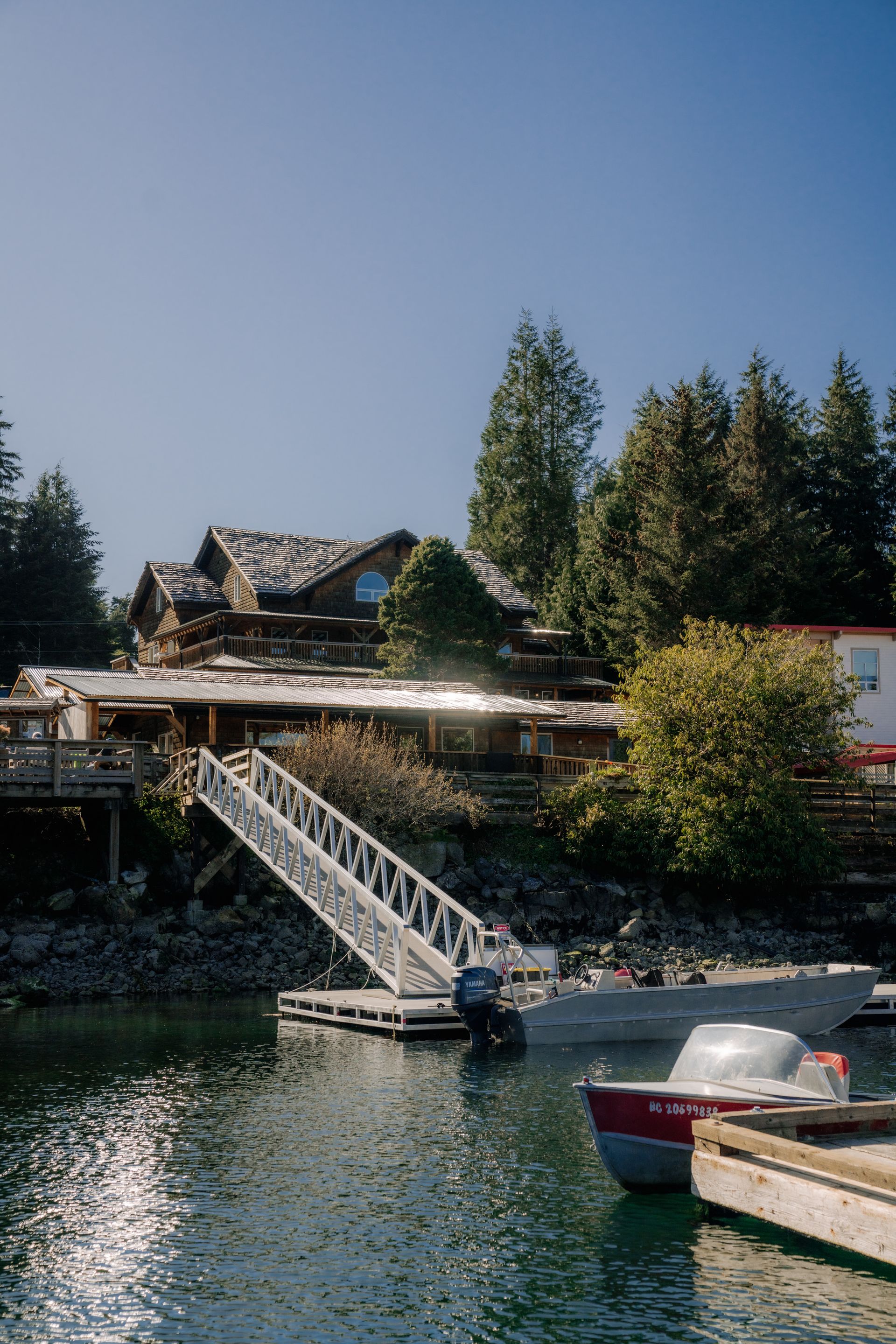 A wooden house sits on a hillside above a dock with a white gangway, a moored boat, and clear blue water.