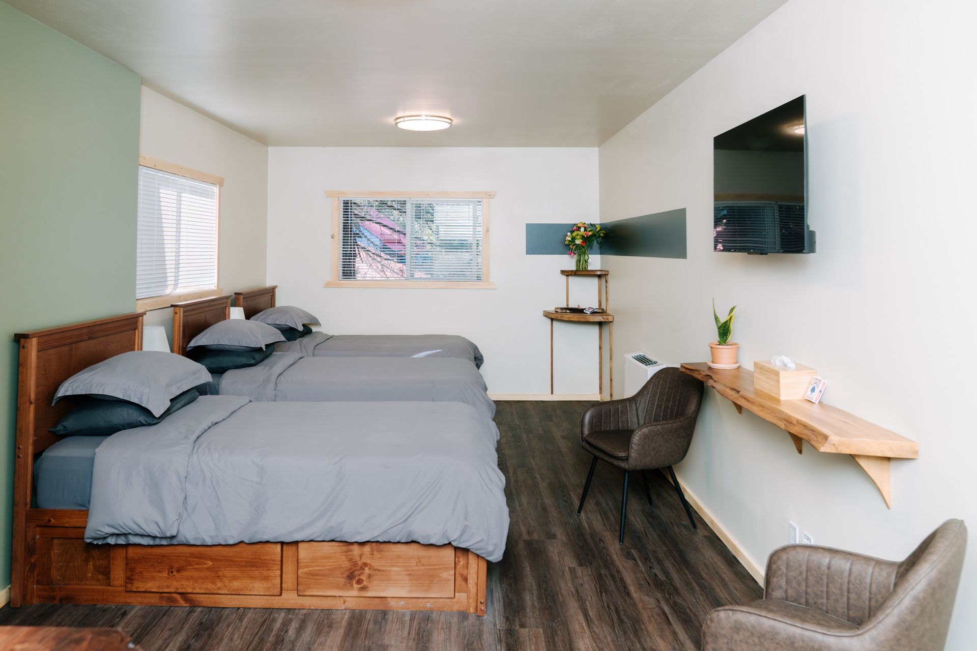 A modern hotel room with two beds with grey bedding, dark wood frames, a wall-mounted TV, two chairs, and wood shelving.