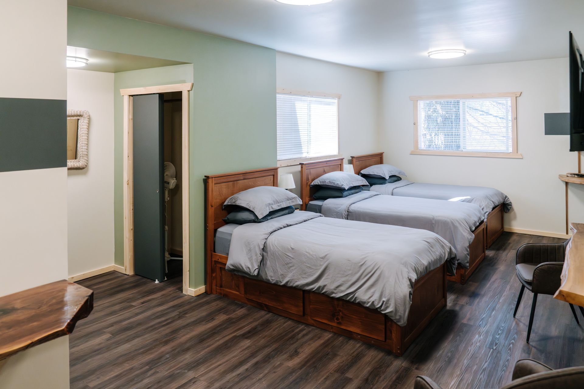 A hotel room with two wooden beds featuring grey bedding, green accent walls, and a view of windows in a neutral space.