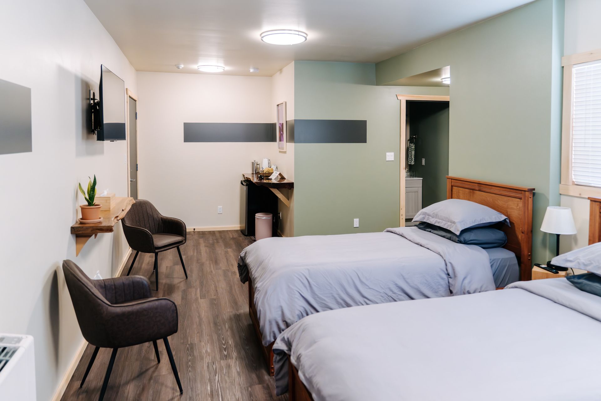 A hotel room with two twin beds, wood headboards, two armchairs, a wall-mounted TV, and a green accent wall.