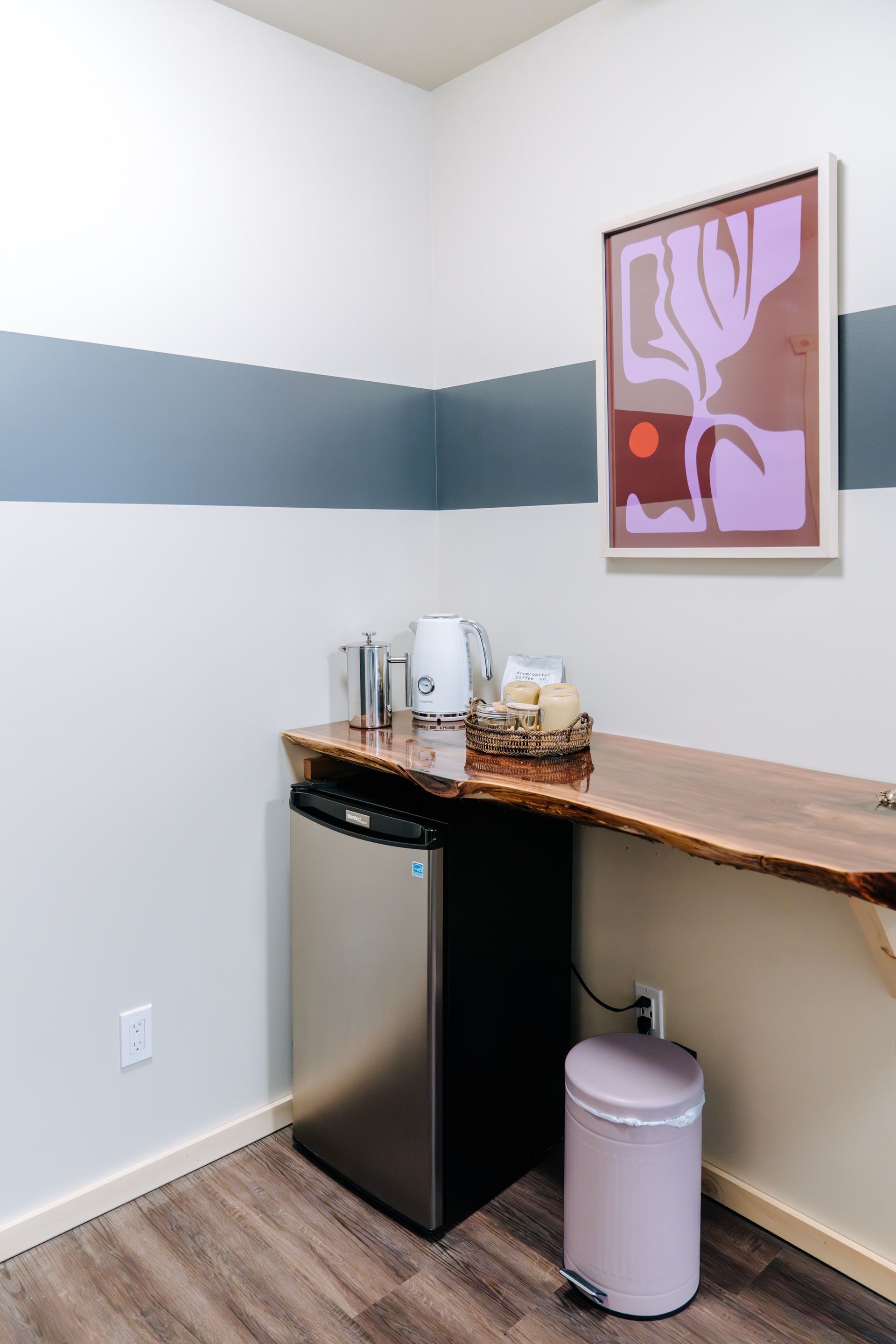 A beverage station with a mini fridge, an electric kettle, and snacks under a wooden shelf against a striped wall.