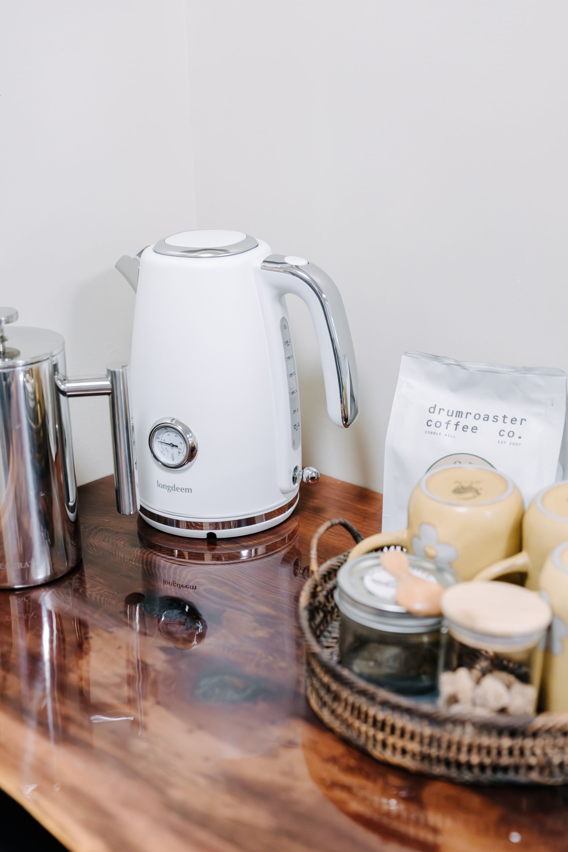 A white kettle, a coffee press, and a basket with coffee supplies and mugs sit on a dark, polished wood surface.