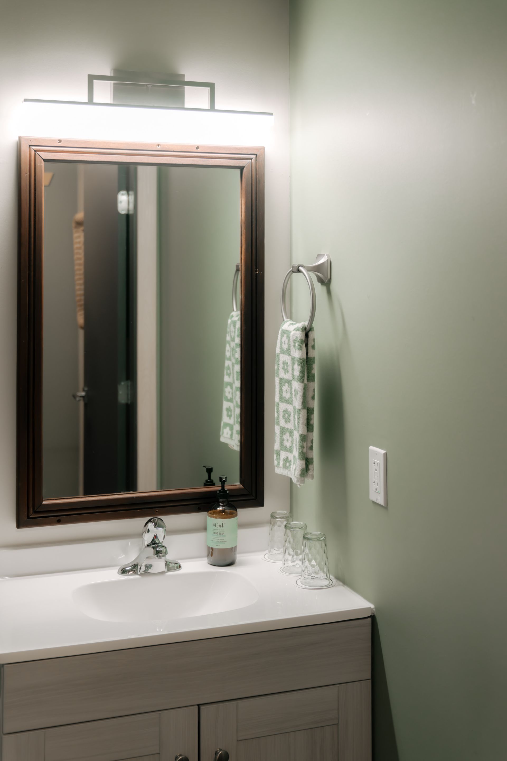A bathroom vanity with a light gray cabinet, white countertop, dark wood-framed mirror, and sage green walls.