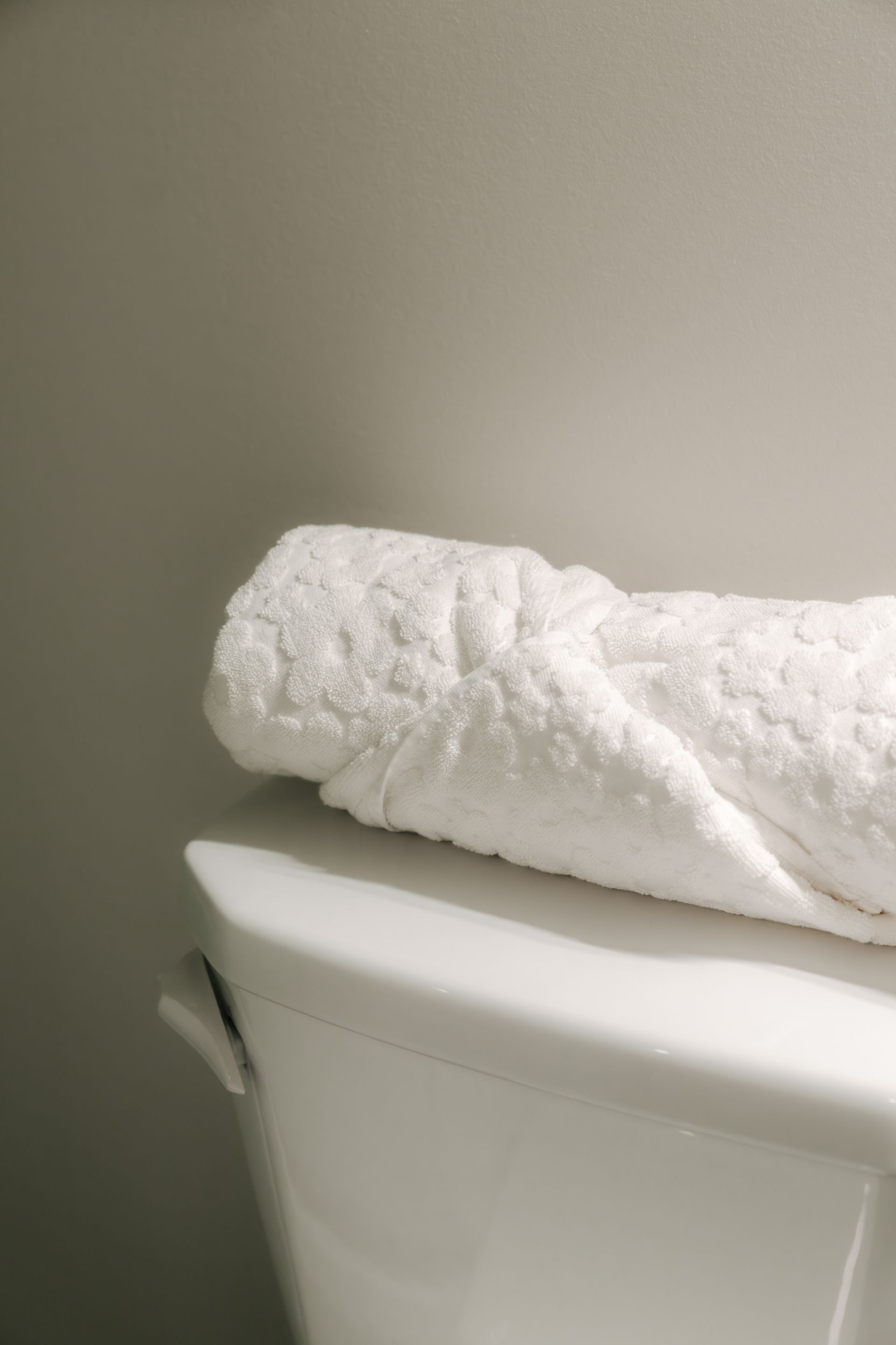 A clean, folded white textured towel sits on top of a white toilet tank against a light gray wall.