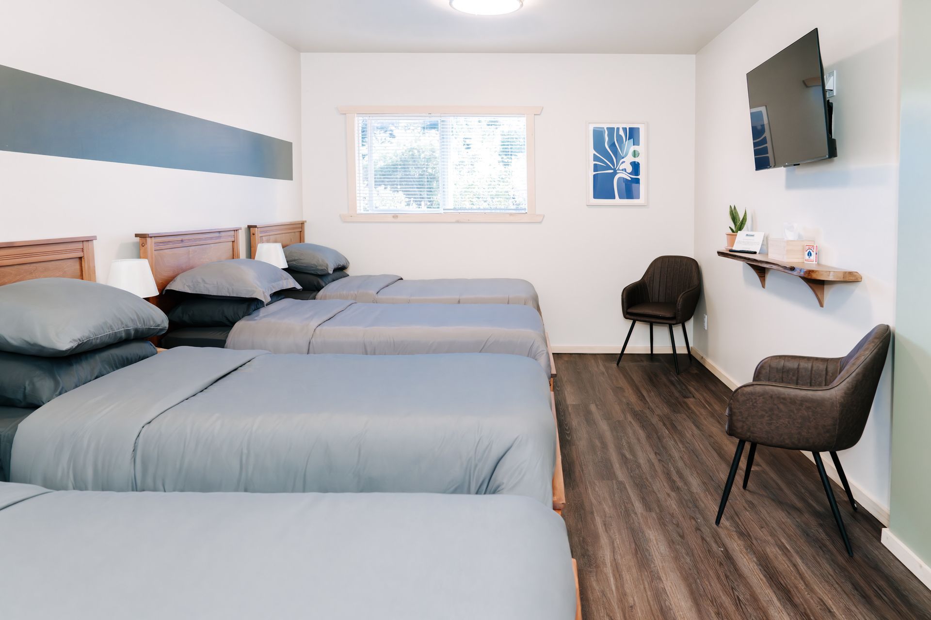 A modern bedroom with three beds featuring grey bedding, a wall-mounted TV, two chairs, and light-colored walls.