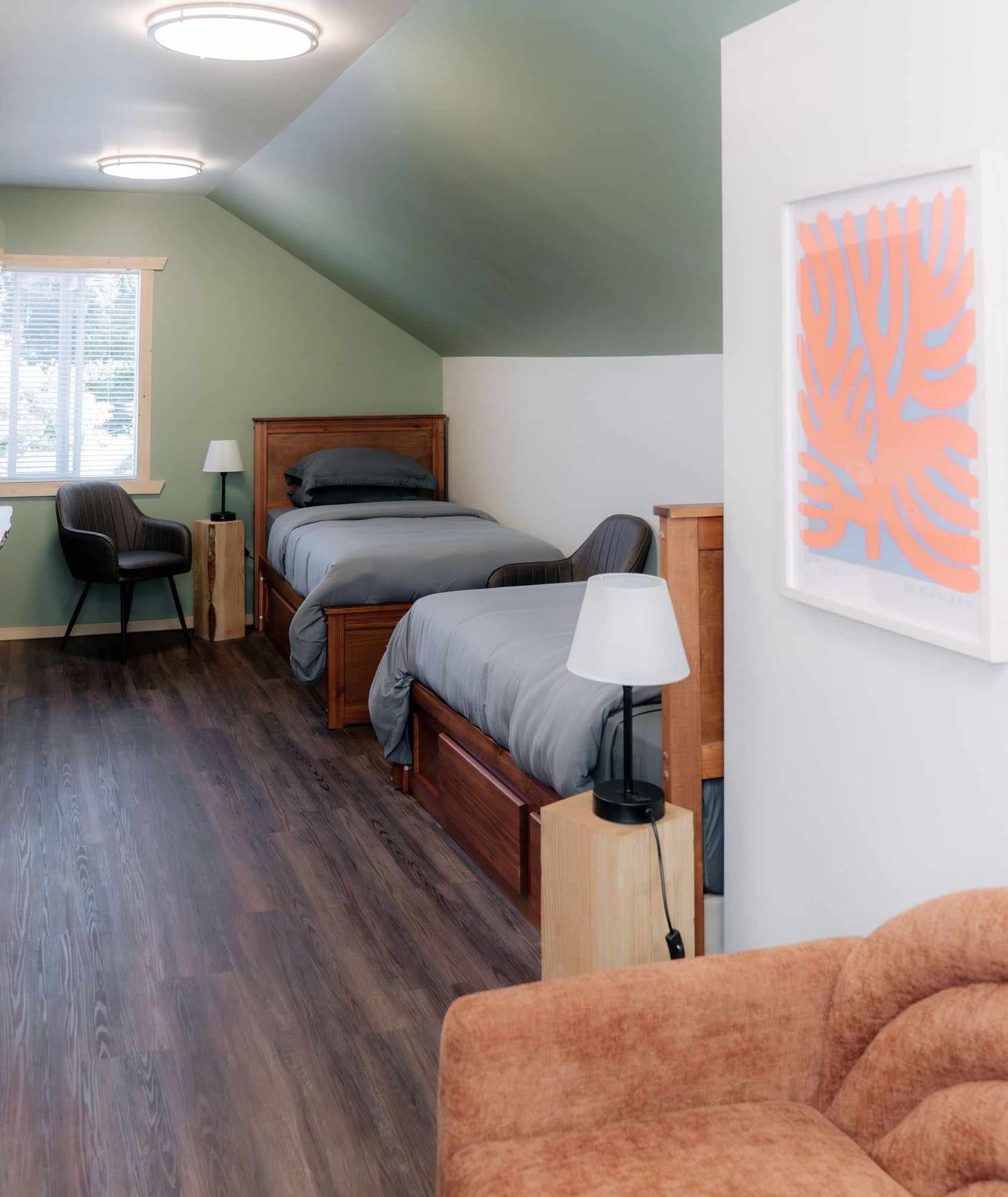 Two beds in a room with a green sloped ceiling, wood floors, a corner armchair, and wall art.