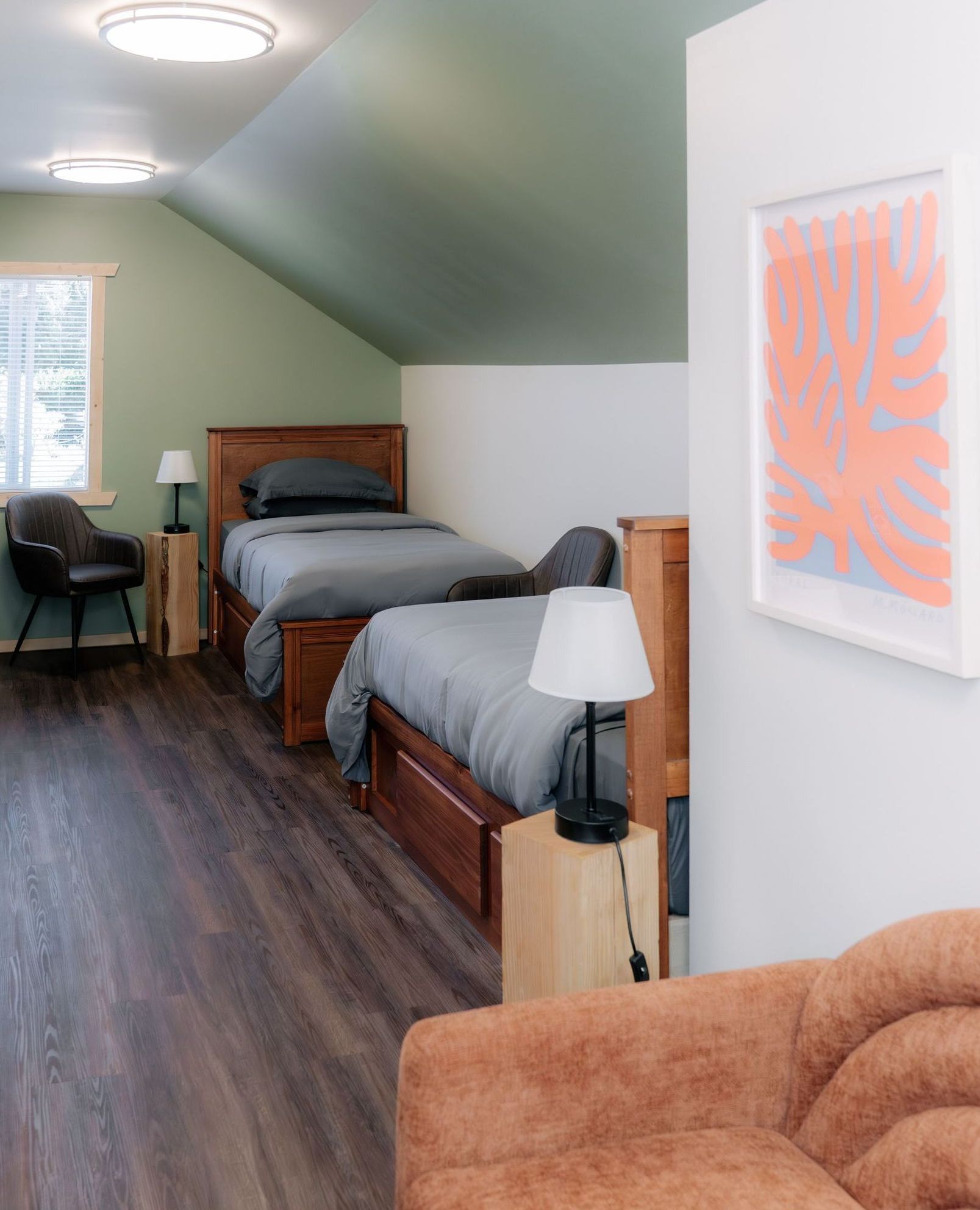 A bedroom with two wooden twin beds, grey bedding, sage green slanted walls, dark floors, and a coral abstract print.