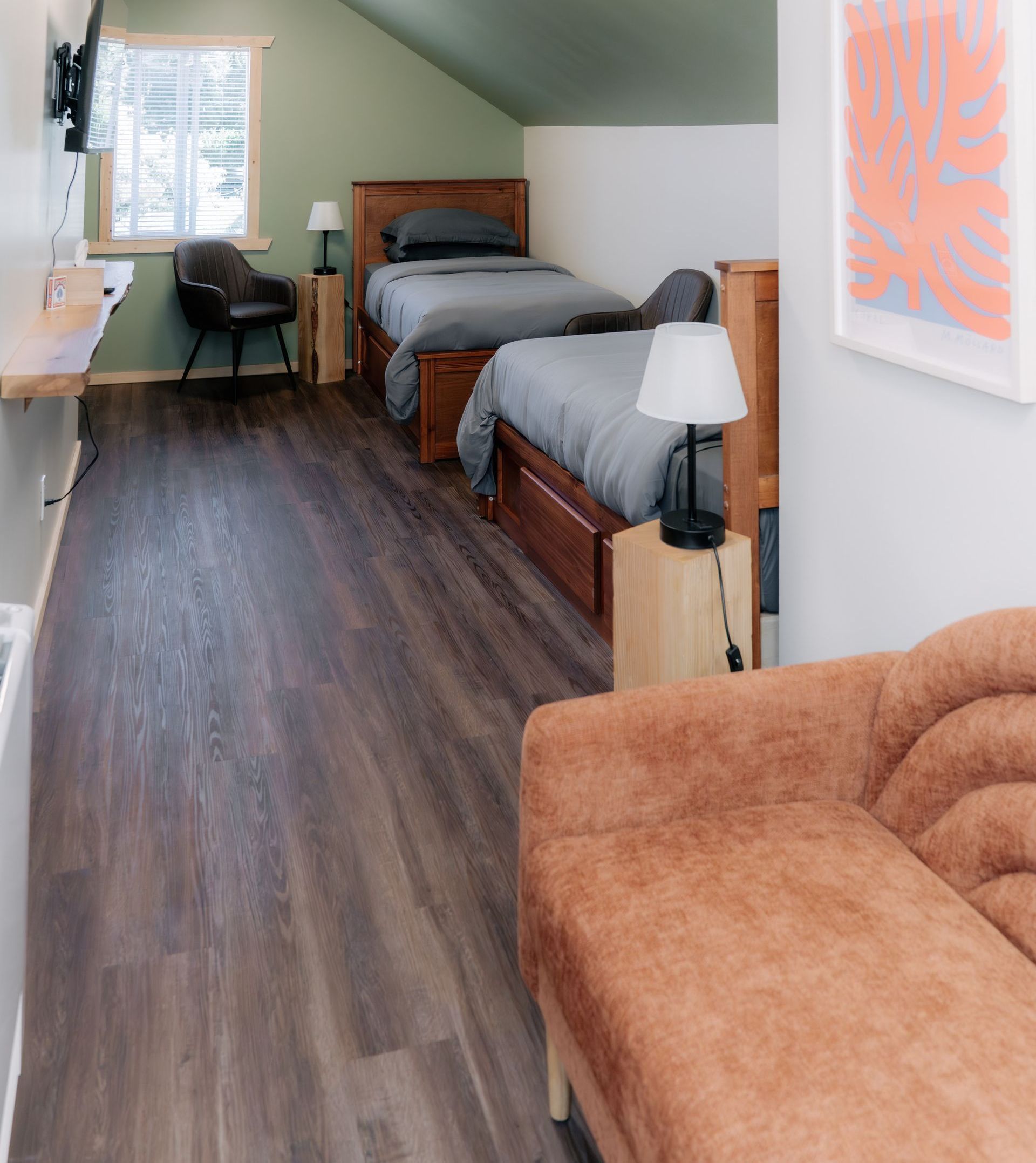 An attic bedroom with two twin wood beds, sage green walls, dark wood floors, and a textured orange couch in the foreground.