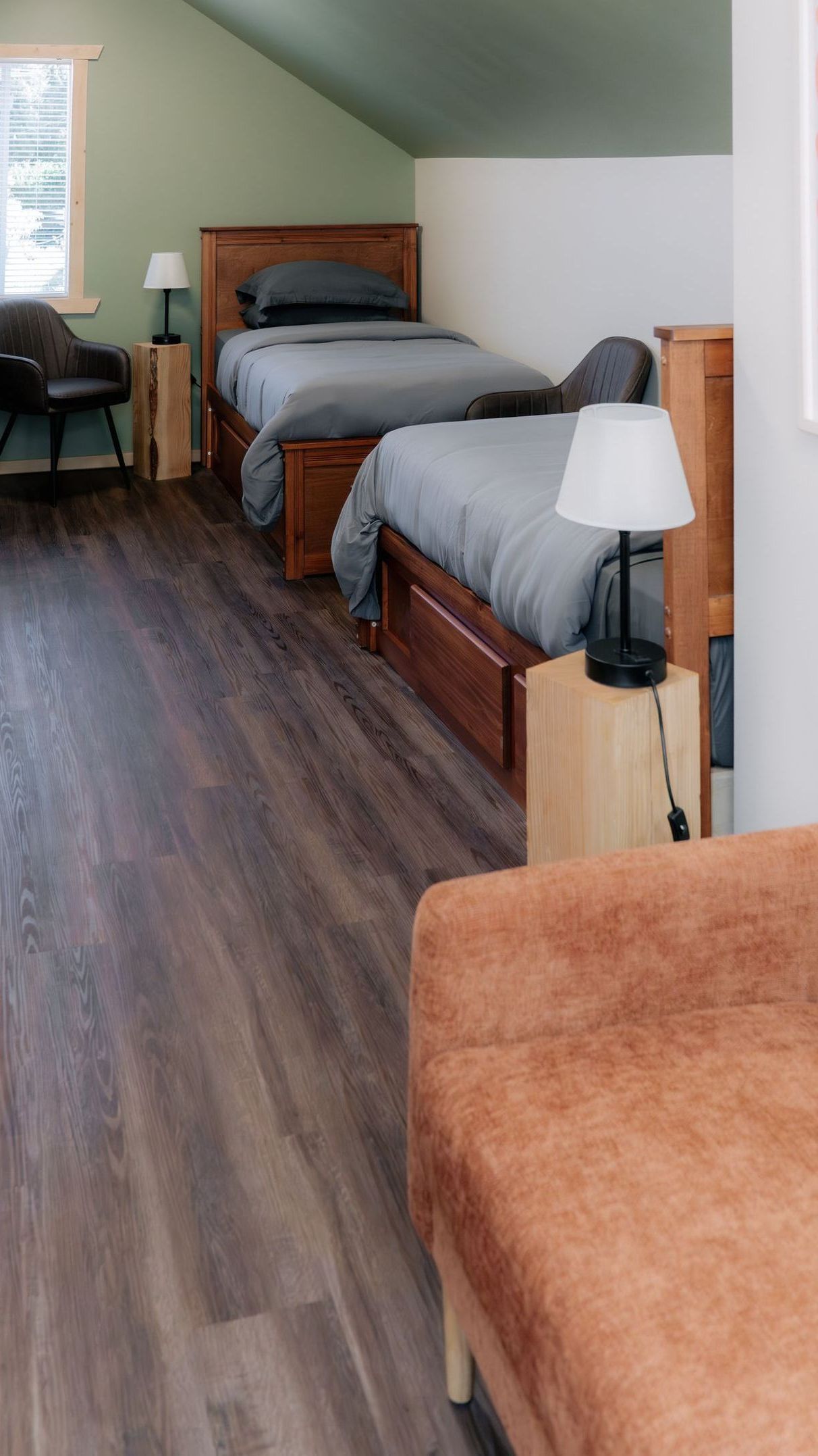 A bedroom with two wooden beds, grey bedding, a side table with a lamp, and a chair near a window on a wood-look floor.
