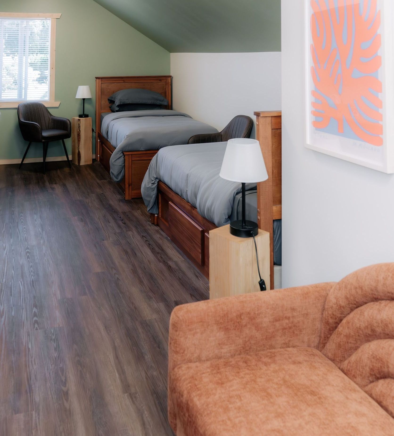 A bedroom with two twin beds, grey bedding, wooden nightstands, a grey armchair, and a tan sofa in the foreground.
