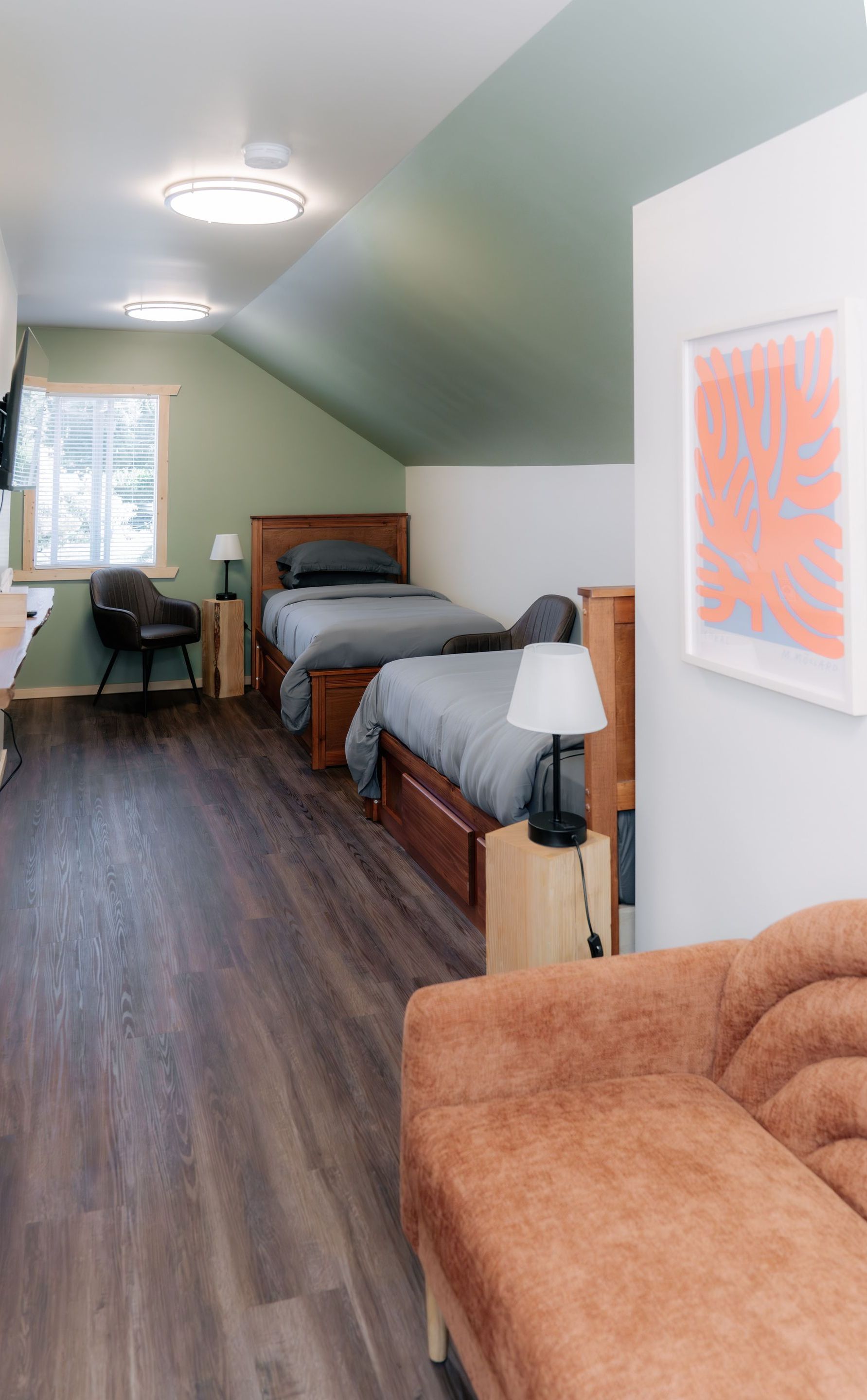 A bedroom with two wooden twin beds, a small seating area, and a beige sofa, featuring green sloped walls and wood floors.