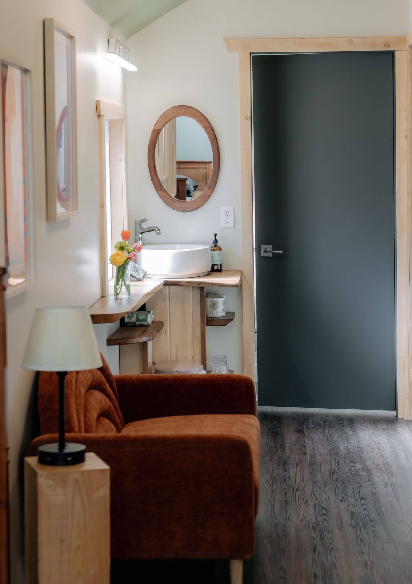 A cozy room with a rust-colored armchair, a lamp, and a wooden shelf vanity with a mirror next to a dark door.