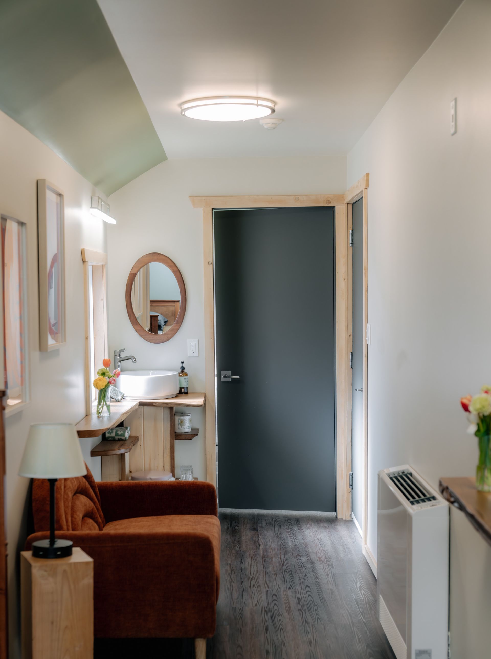 A cozy hallway with a dark brown armchair, a wooden vanity with a round mirror, and a dark grey door.