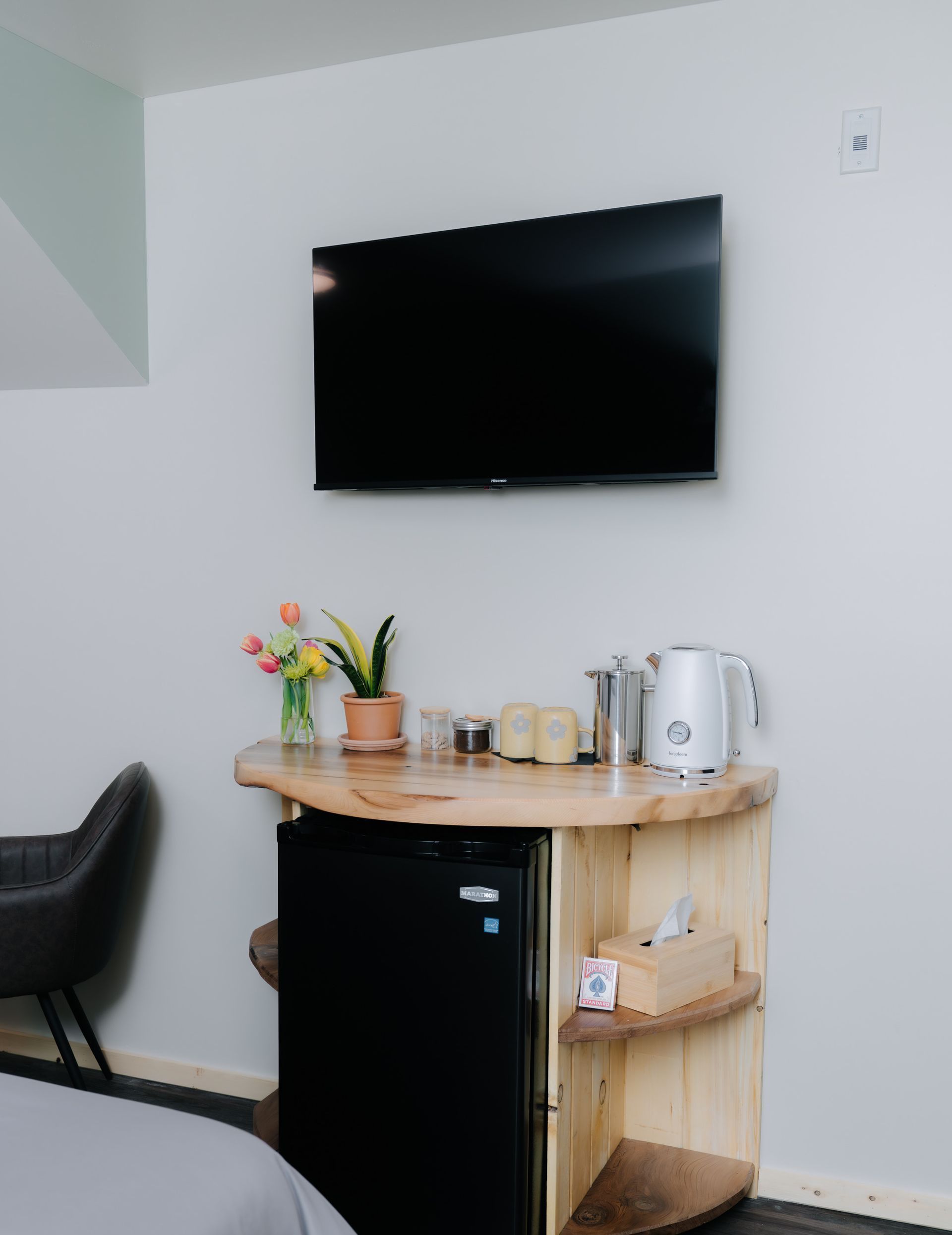 A wall-mounted TV above a light wood counter holding a small fridge, kettle, and potted plant in a modern room.