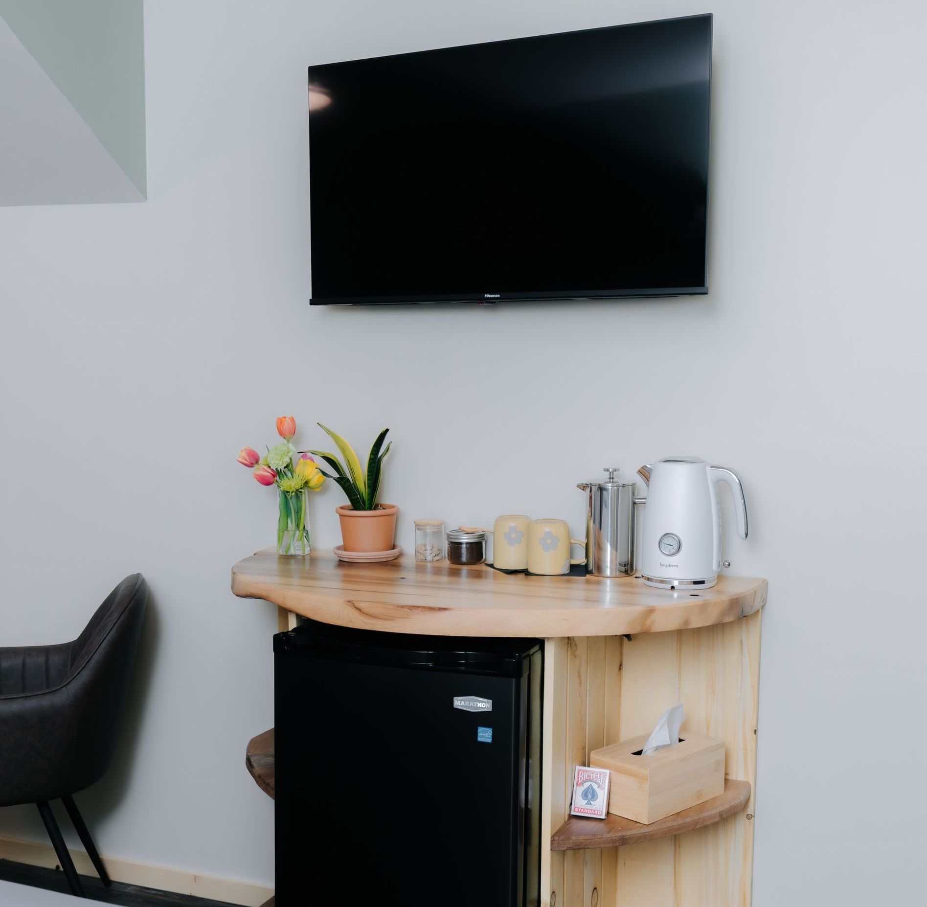 A TV mounted above a curved wooden counter with a mini-fridge, tea kettle, and a small vase of flowers.
