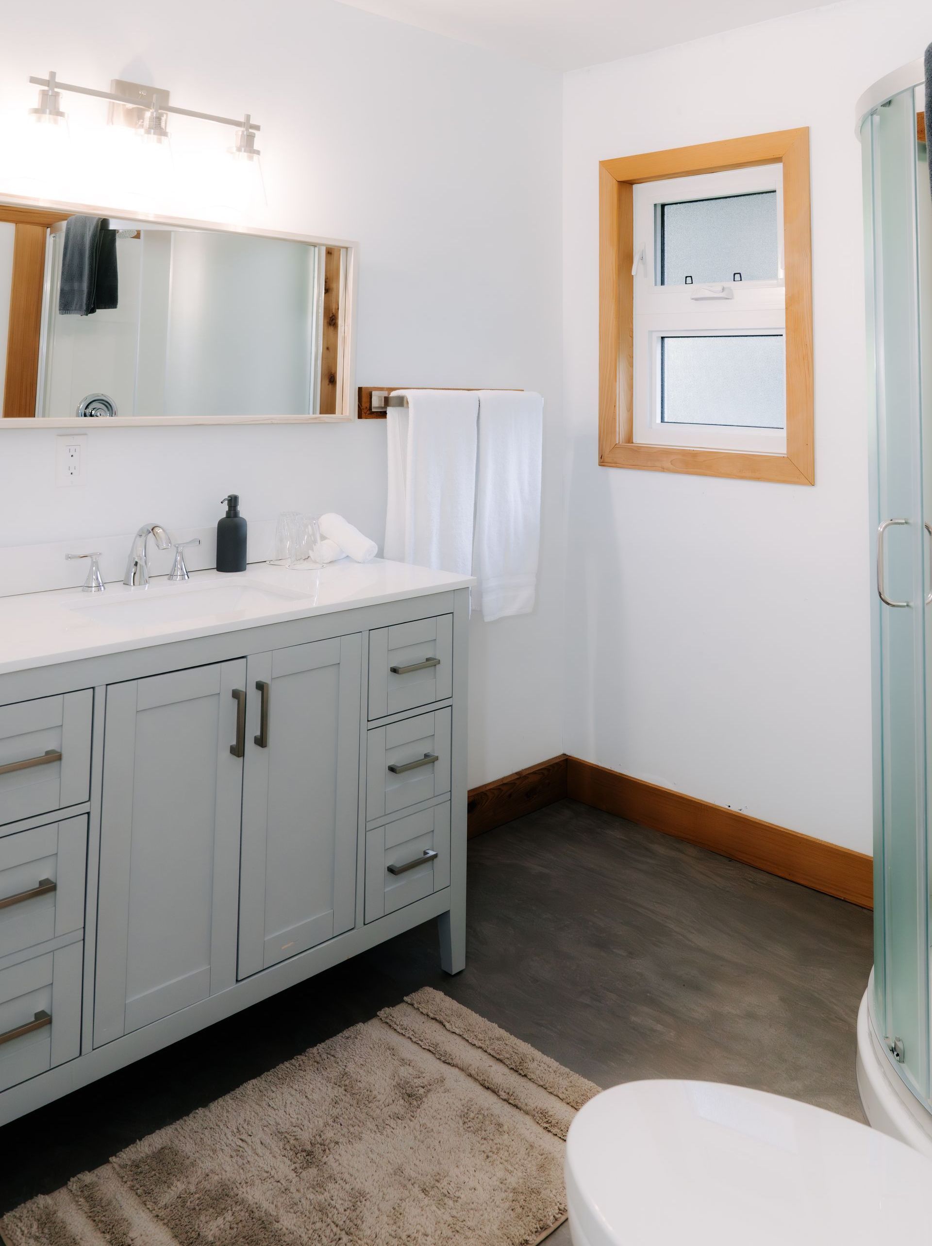 A light gray bathroom vanity with white countertops, a mirror, and a window in a white-walled room with dark flooring.