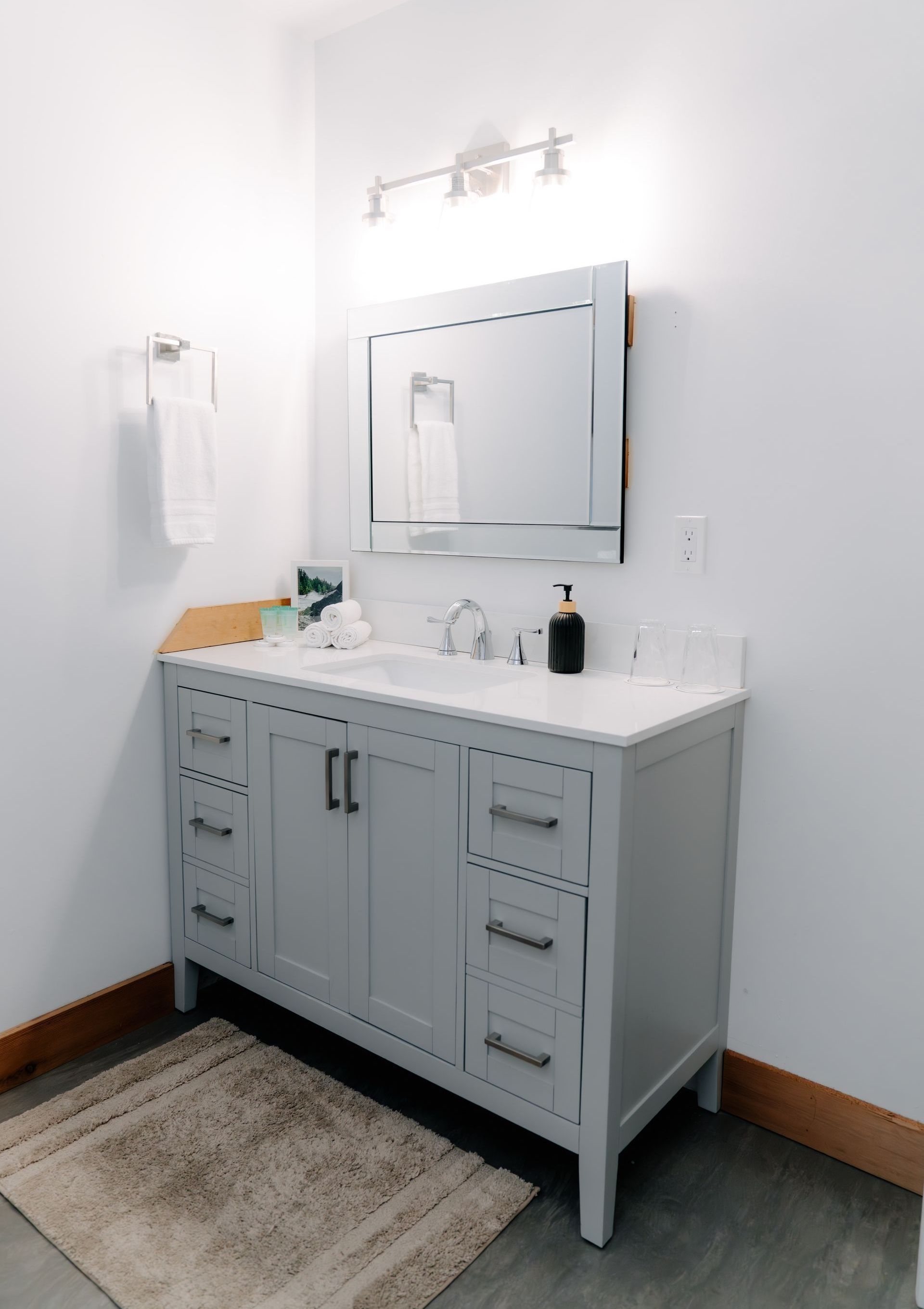 A gray bathroom vanity with a white countertop, mirror, and light fixture, set against white walls on a gray floor.