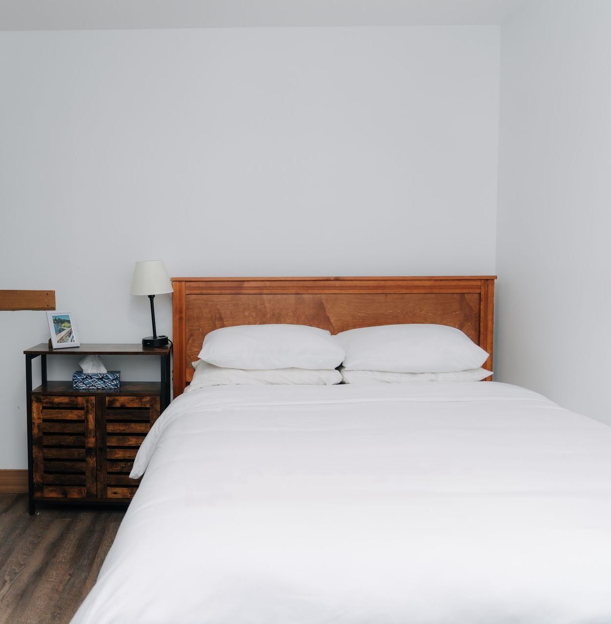 A white bed with a wooden headboard next to a dark wood bedside table with a lamp in a minimalist bedroom.