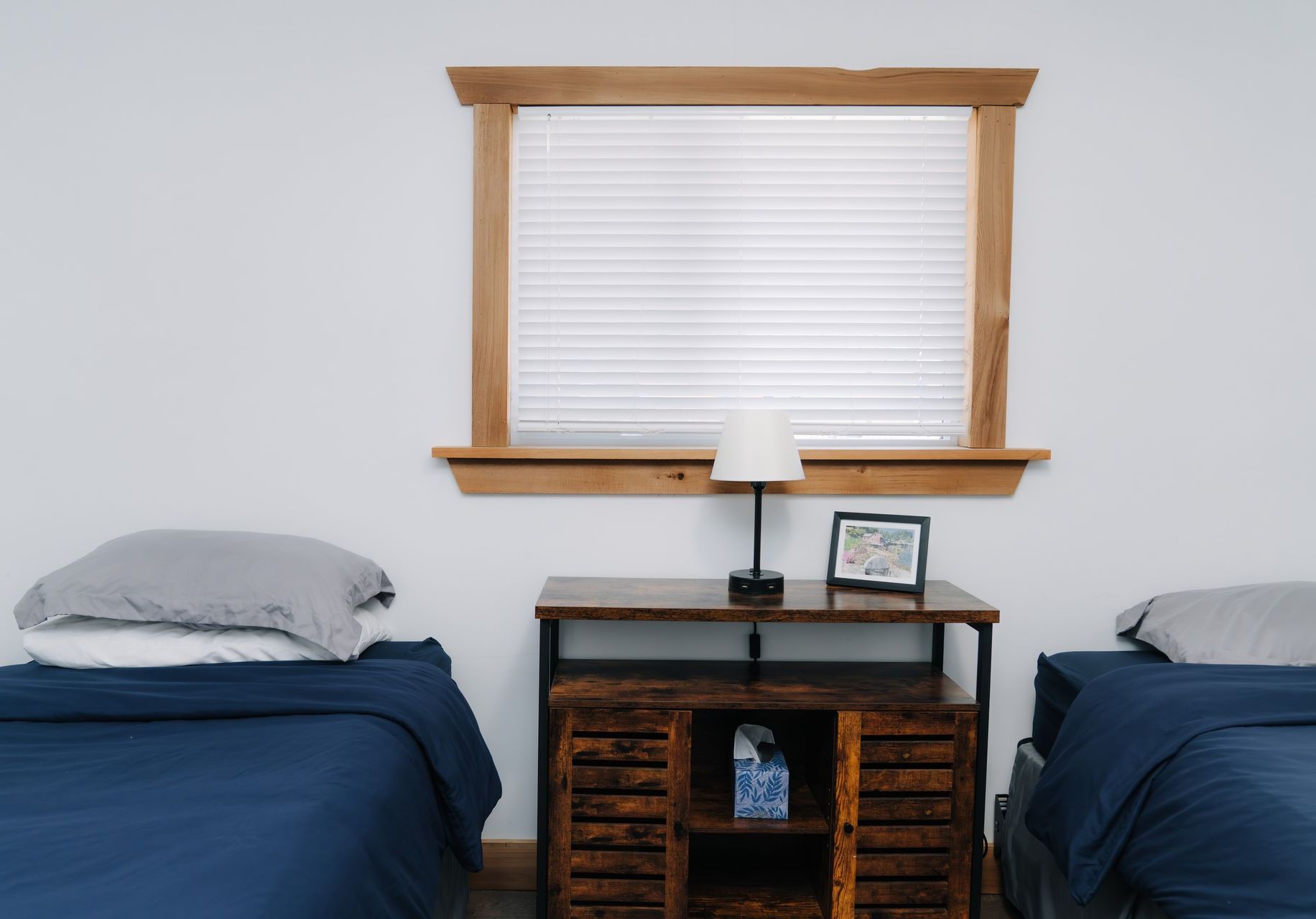 Two beds with blue bedding and grey pillows, separated by a wooden console table featuring a lamp and a small picture.