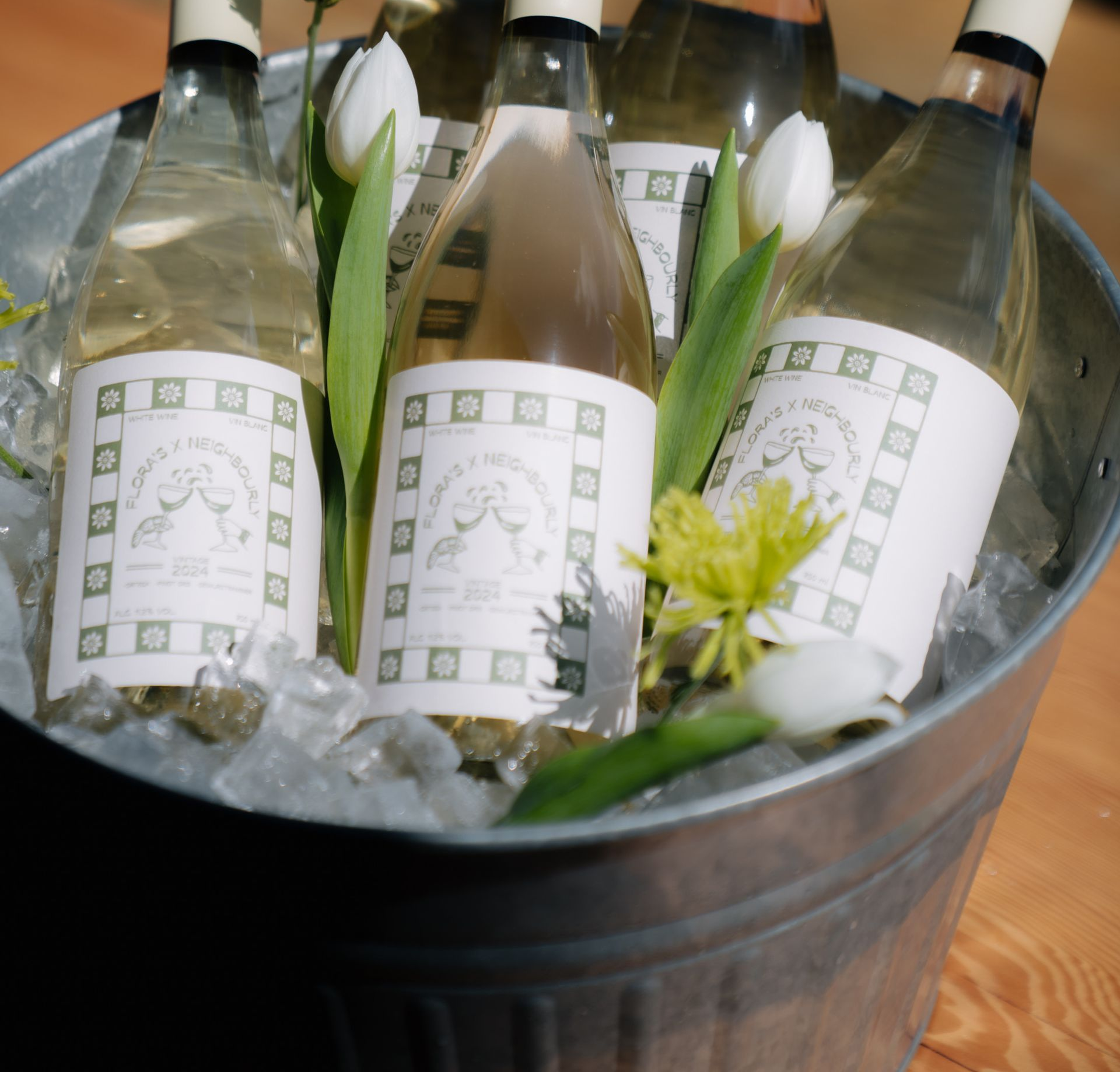 Four wine bottles with matching custom labels in a metal bucket filled with ice and white tulips.